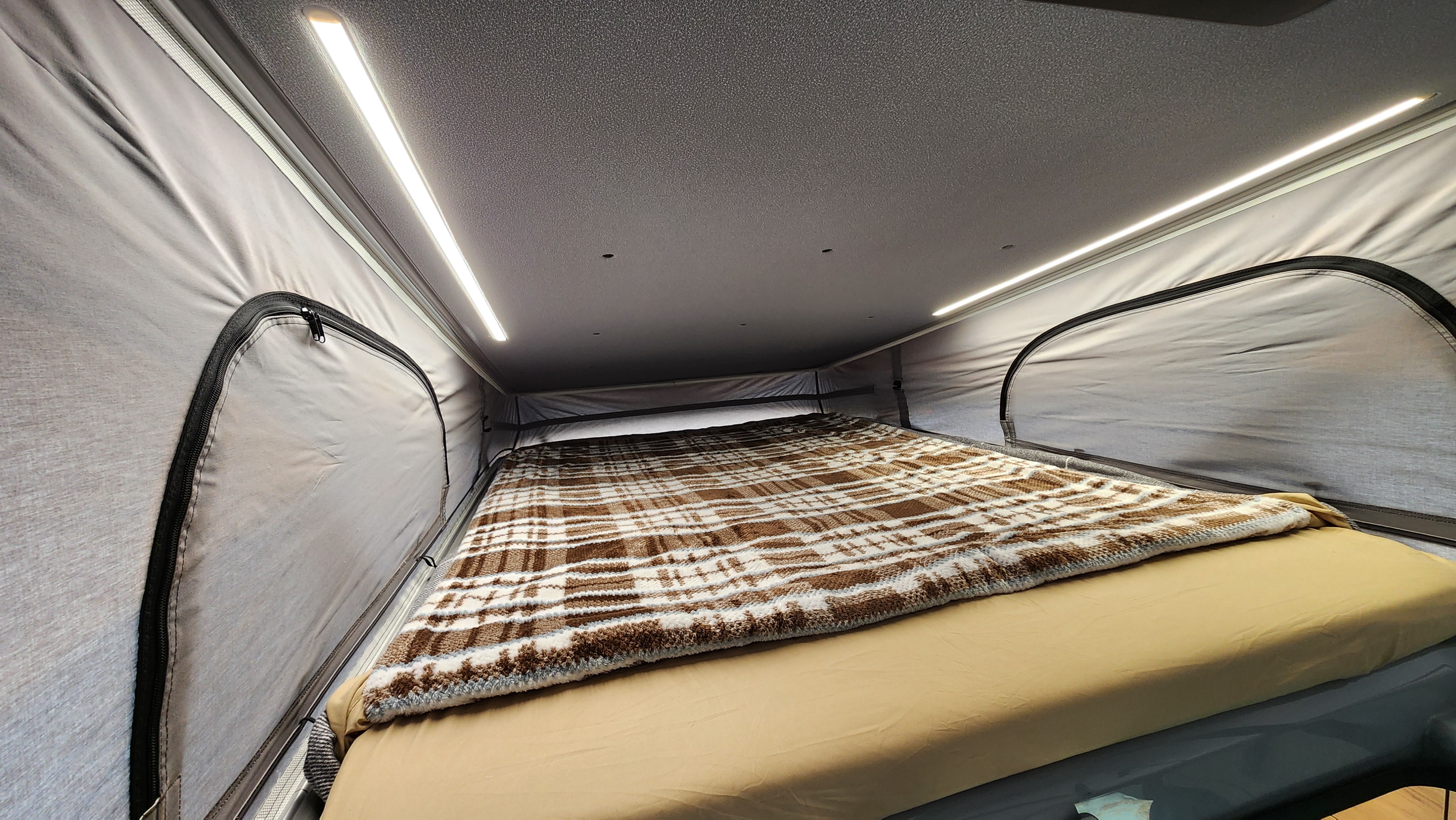 RV rental's cozy pop-top bed features a brown plaid blanket, beige sheets, and bright LED lighting for comfortable camping.. Airstream Rangeline 2024