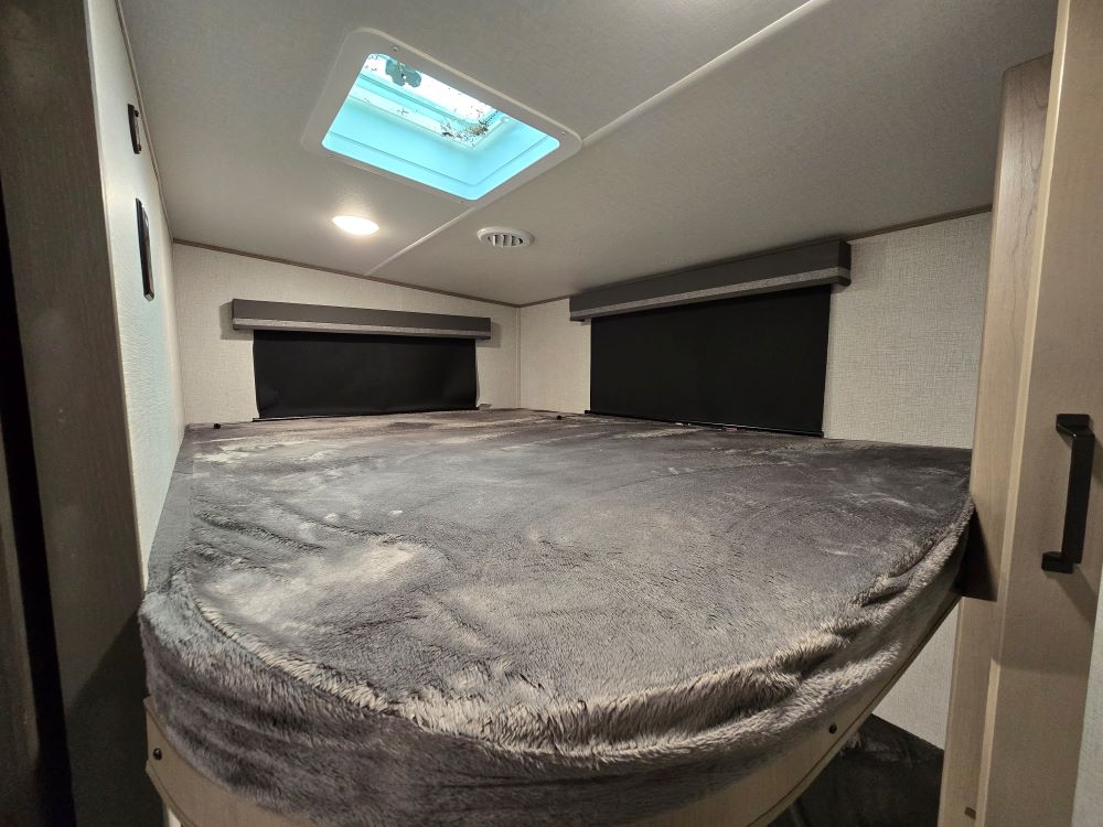 RV rental interior: large bed with a soft grey cover, overhead skylight, and privacy blinds.. Grand Design Reflection 150 2023