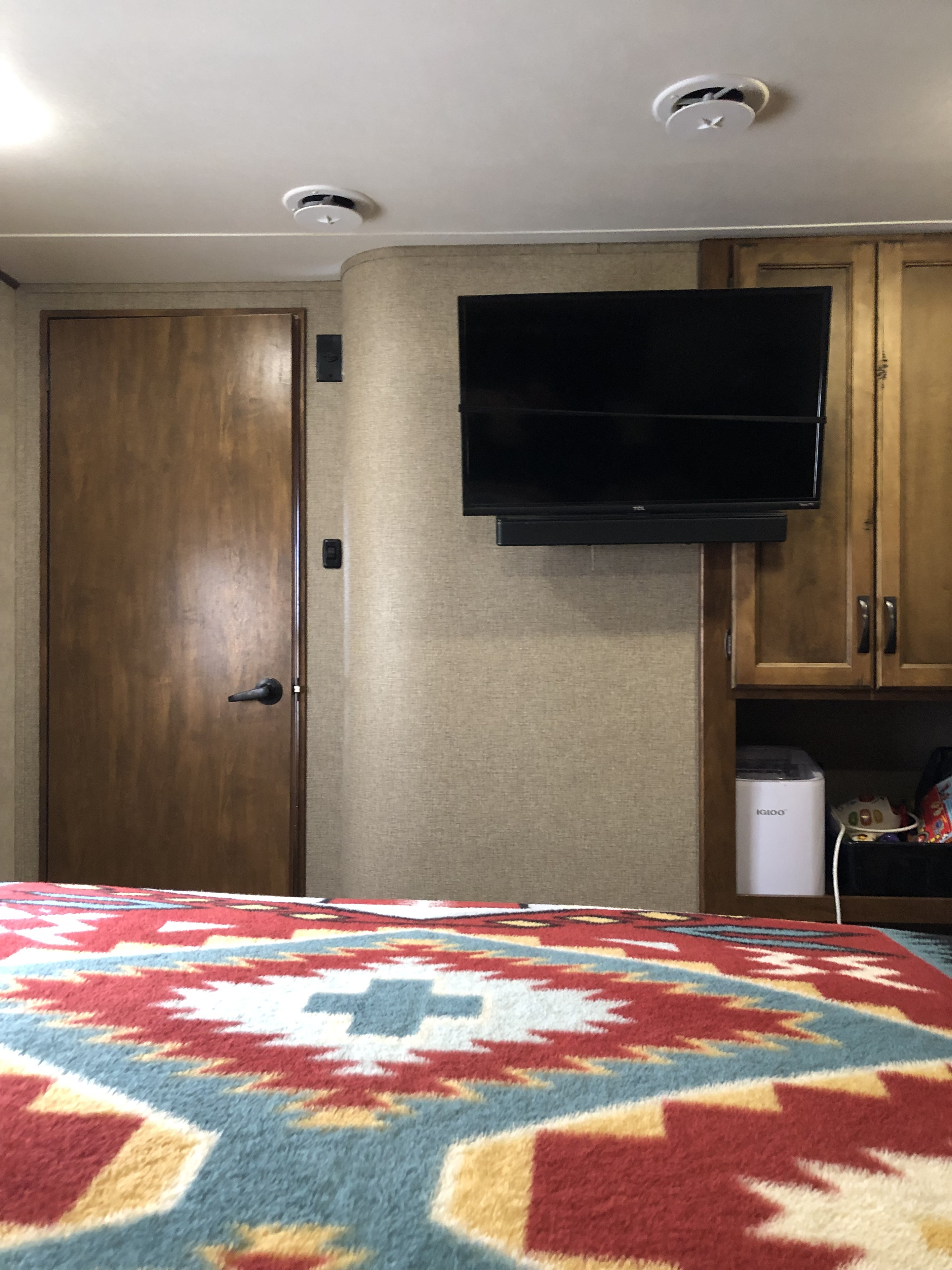 Inside a comfy RV rental bedroom: vibrant patterned blanket, wall-mounted TV, soundbar, and Igloo ice maker.. Keystone Carbon 2015