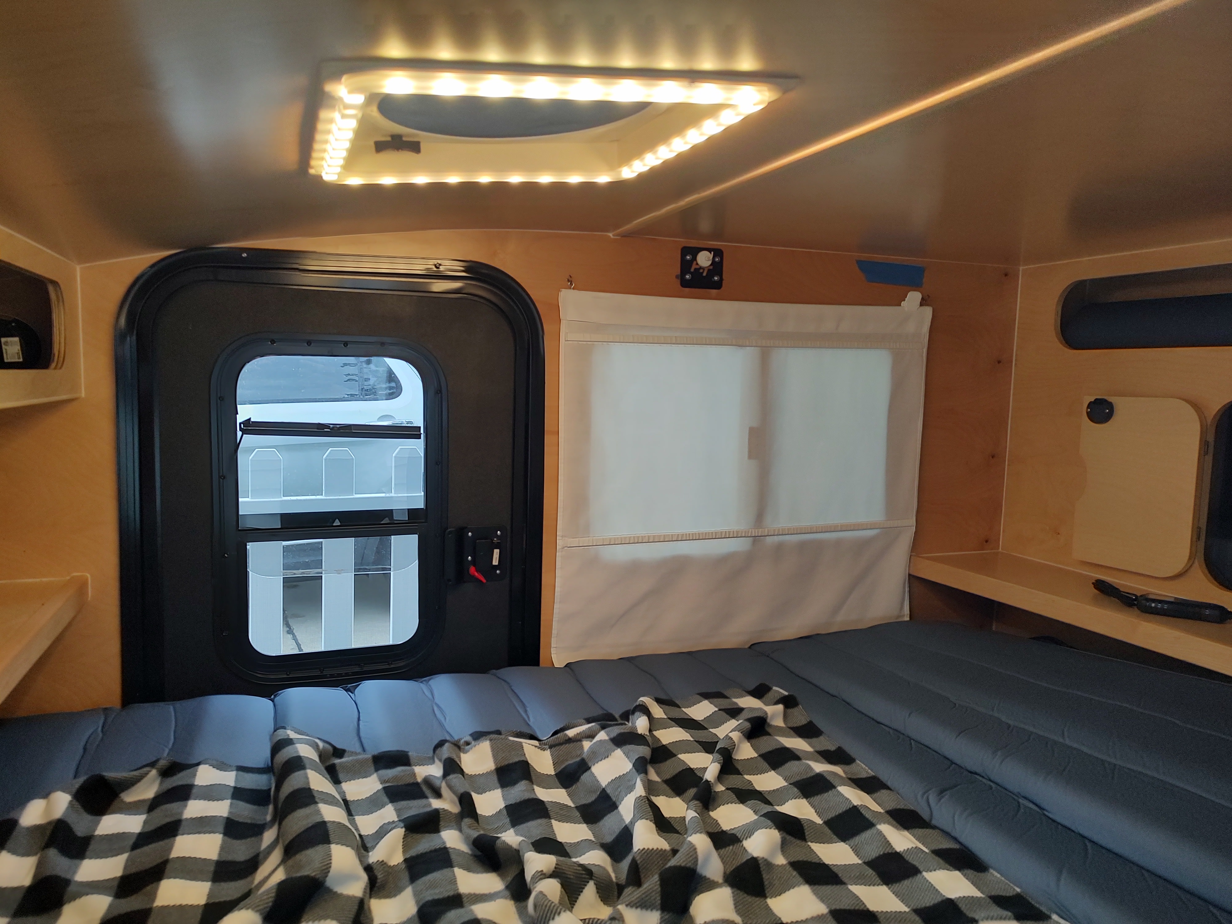 Compact RV rental interior featuring a comfortable bed with plaid blanket, a door, window with shade, and overhead LED lighting.. Hiker Trailer 5x9 MR Delux 2022