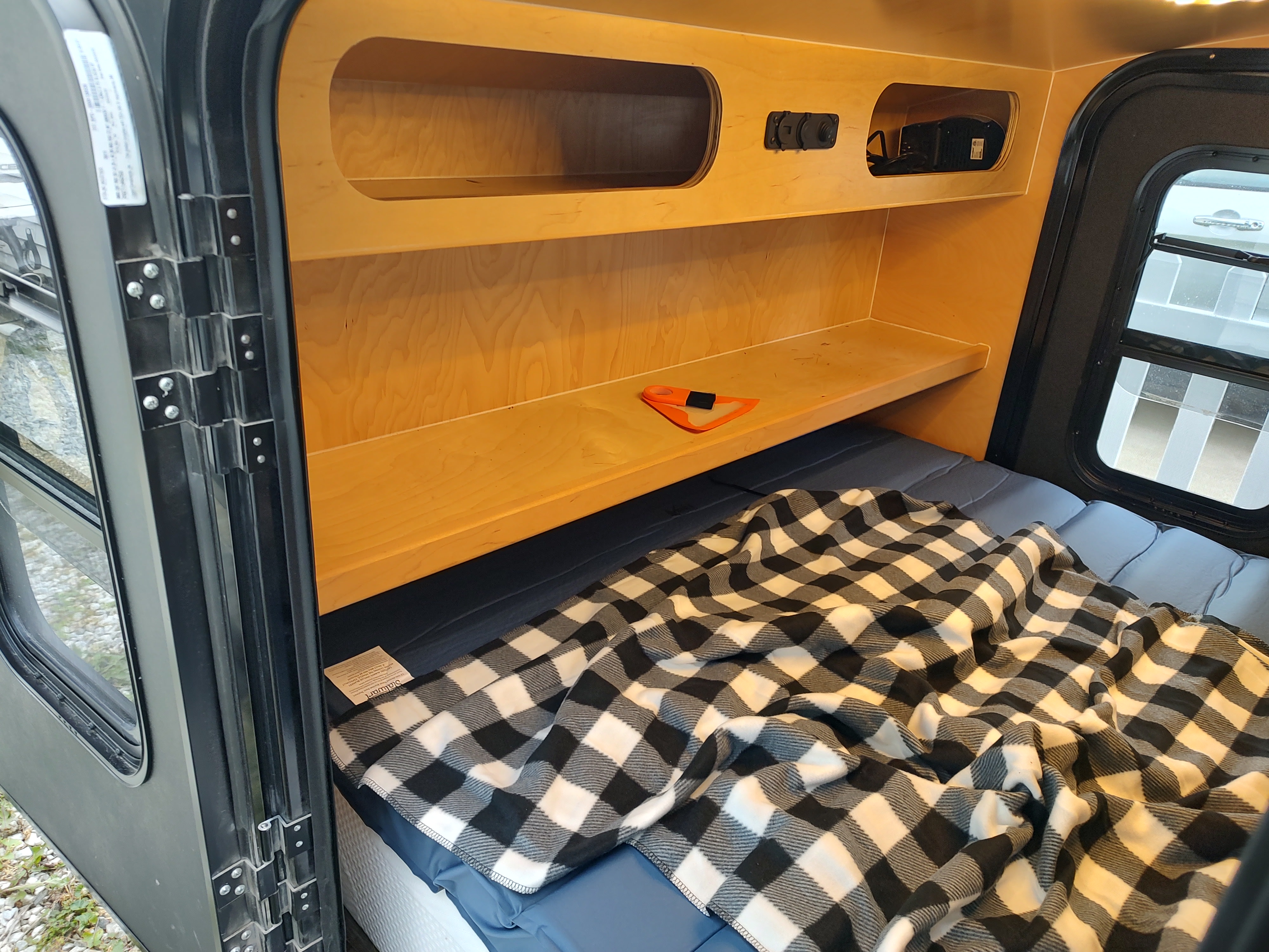 Explore this RV rental's comfy interior: a bed with checkered blanket, built-in wooden storage, and a large window.. Hiker Trailer 5x9 MR Delux 2022