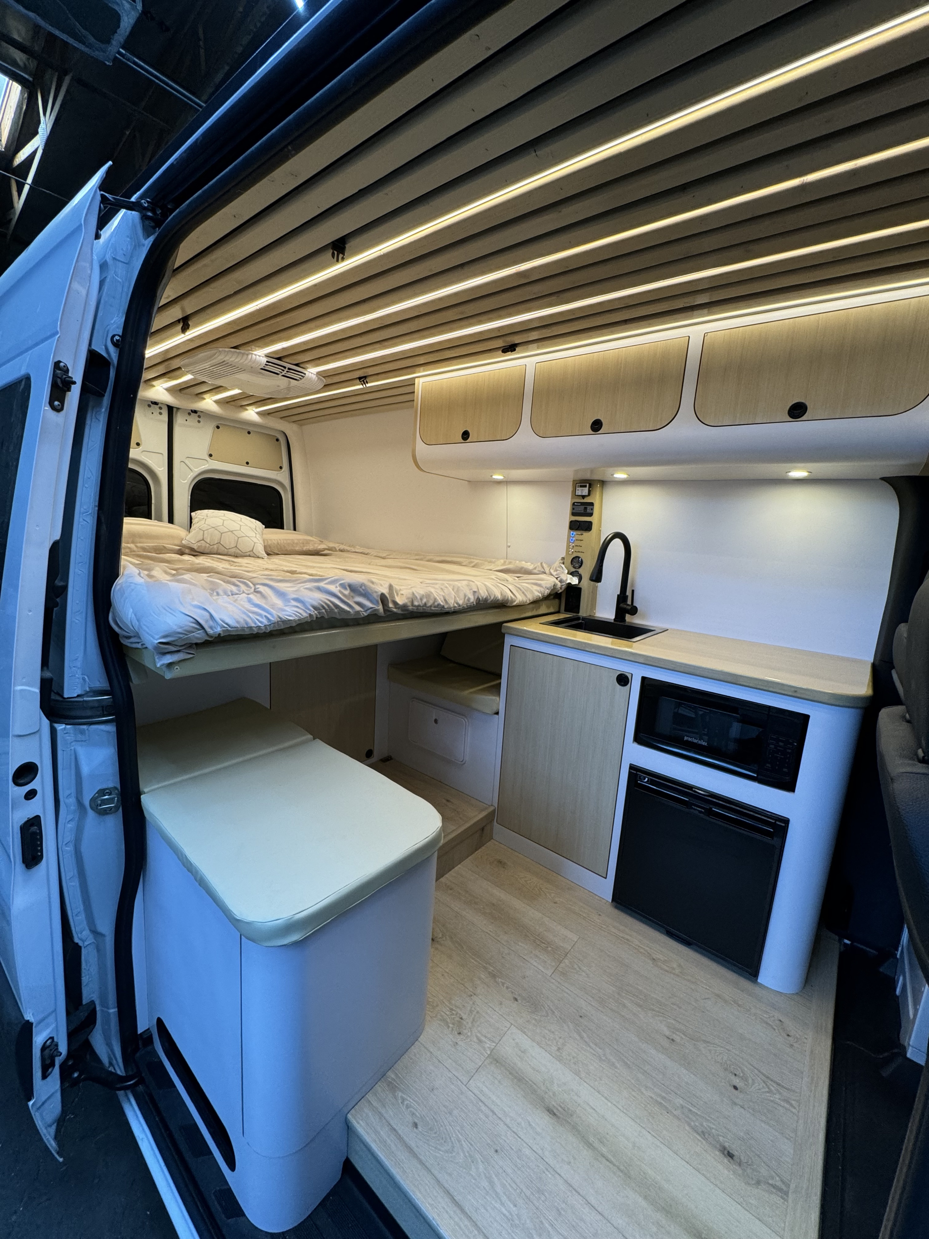 Modern RV rental interior with a raised bed, compact kitchen (microwave, fridge), overhead cabinets, and warm ceiling lights.. Mercedes-Benz Sprinter 2022