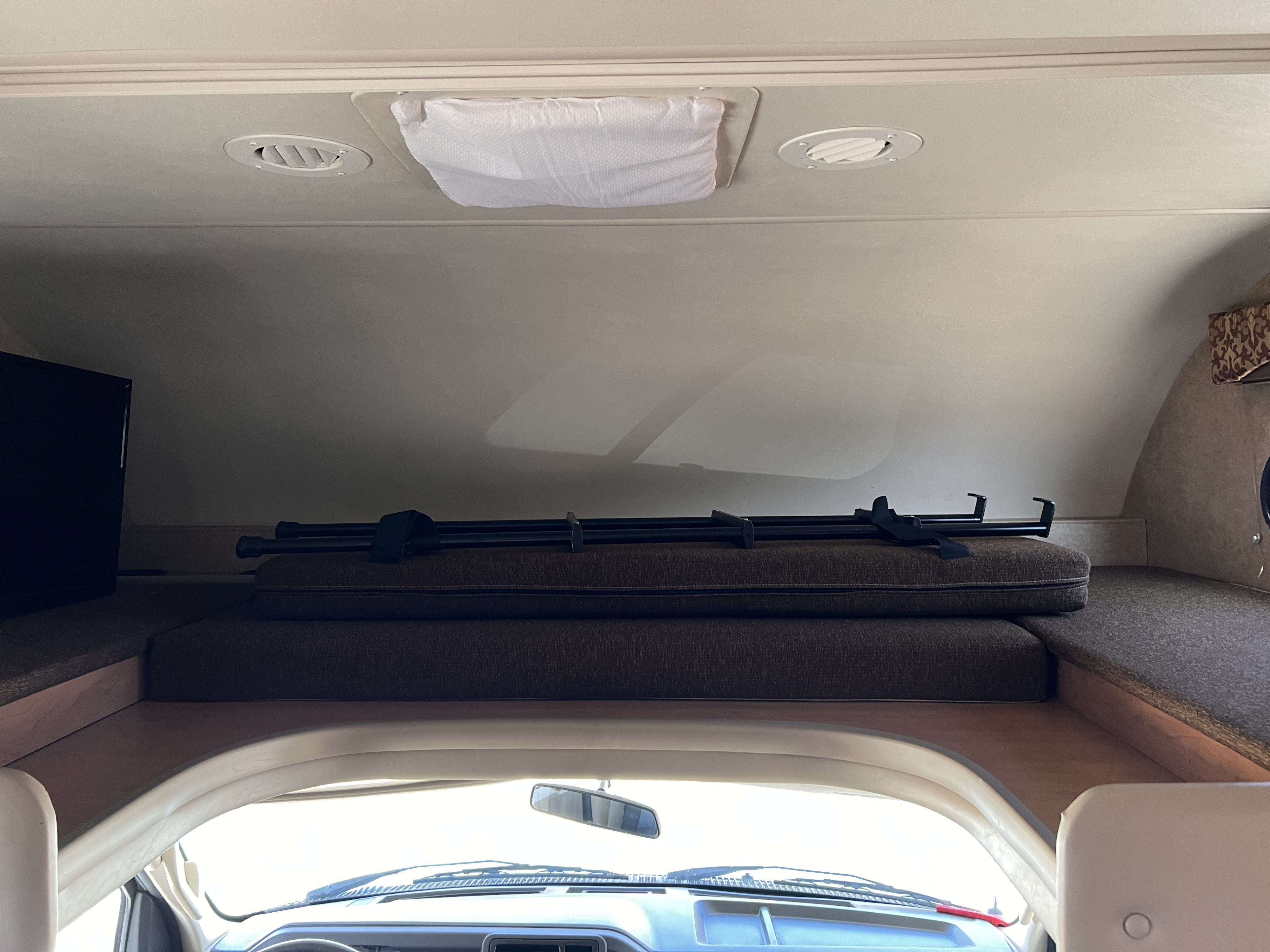 RV rental interior: over-cab bunk with mattress & support bars. Ceiling vents, a covered vent, & TV. View from the driver's seat.. Thor Motor Coach Freedom Elite 2017