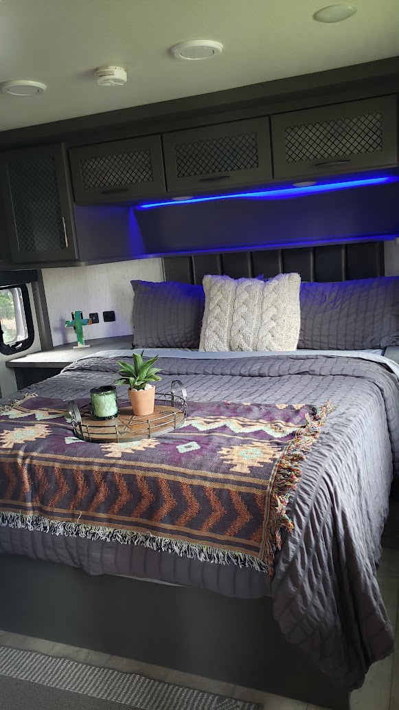 Cozy RV rental bedroom with gray bedding, a vibrant patterned throw, and blue accent lighting. Note the overhead cabinets.. Grand Design Momentum Toy Hauler 2021