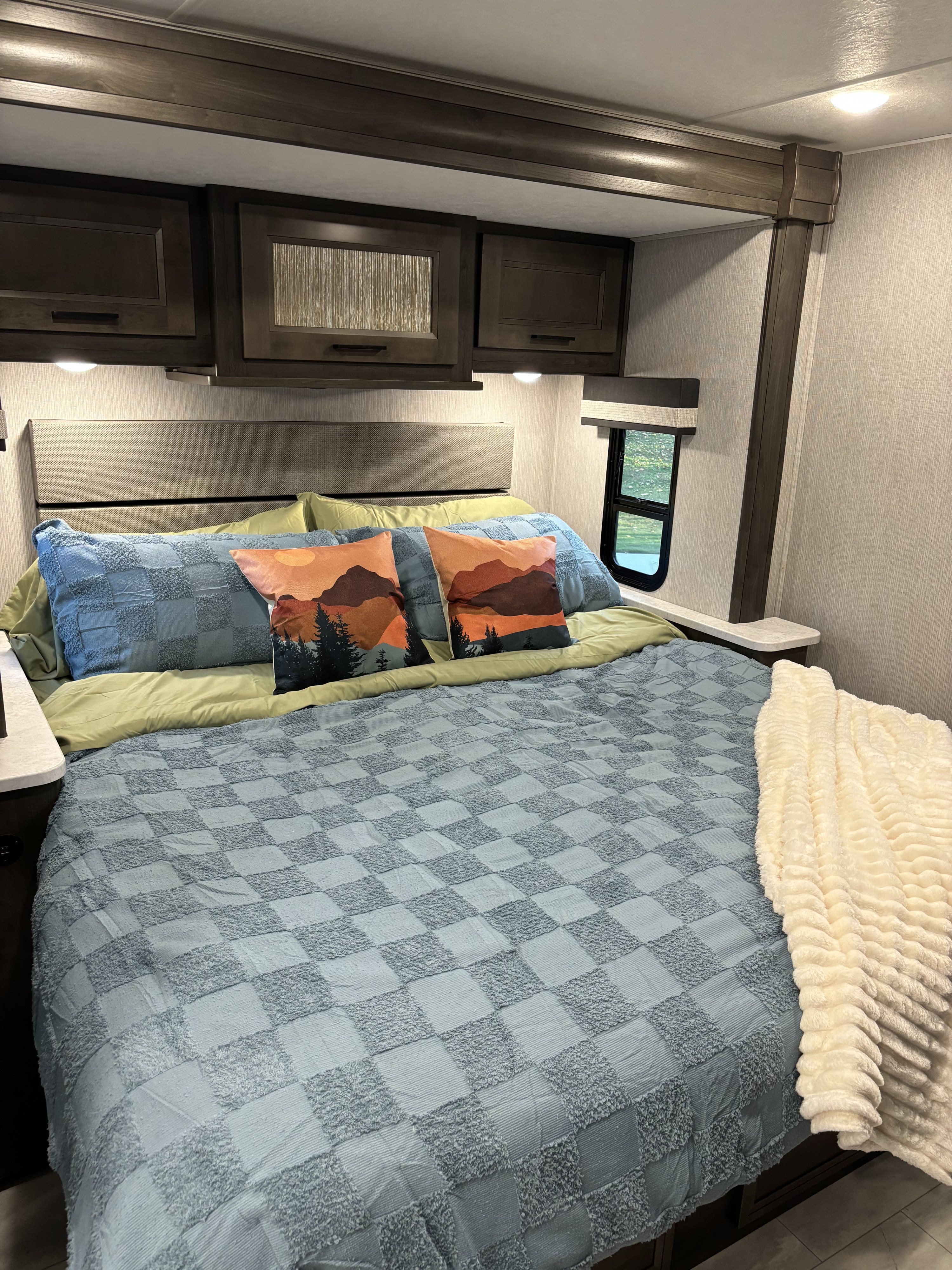 Cozy RV rental bedroom with checkered bedspread, mountain pillows, overhead cabinets, and a window.. Winnebago Coachmen 35ES Class A RV 2024