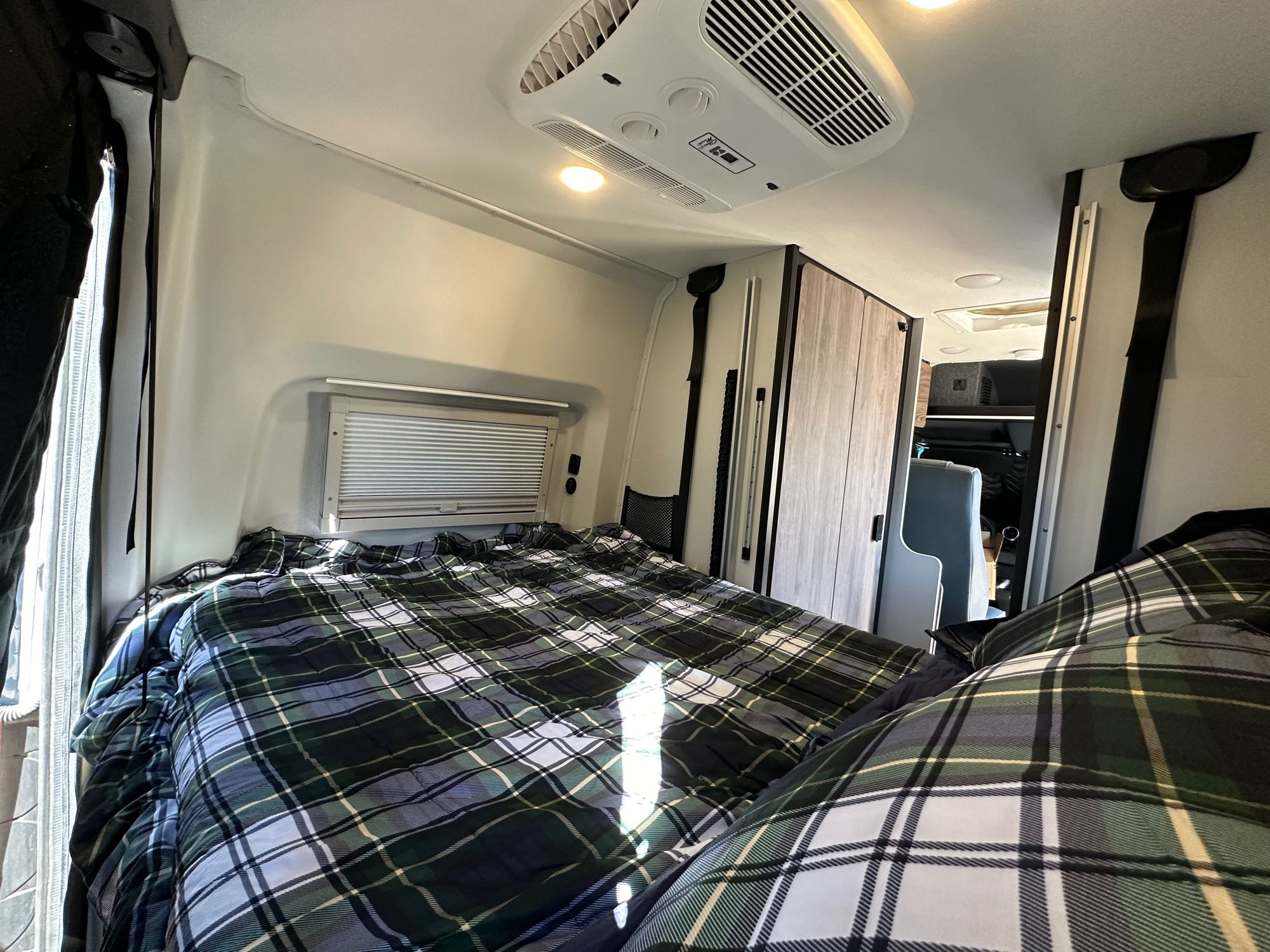 Cozy plaid bed in an RV rental, filling the frame. Features ceiling AC, window, and wood-paneled storage.. Tiffin GH1 2023