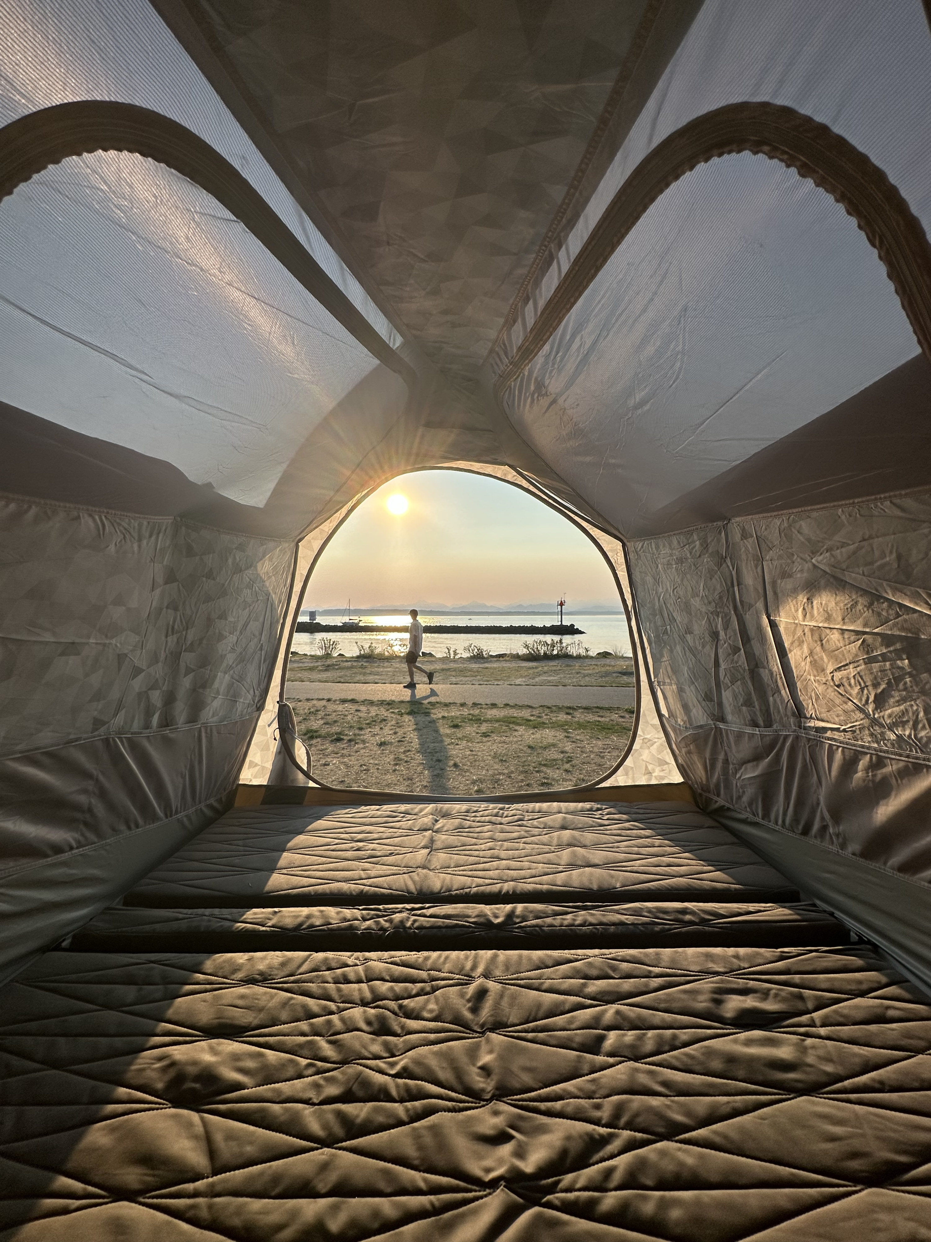From an RV rental, a comfy quilted bed frames a golden sunset over water with a person walking outside.. Tesla Cybertruck 2024