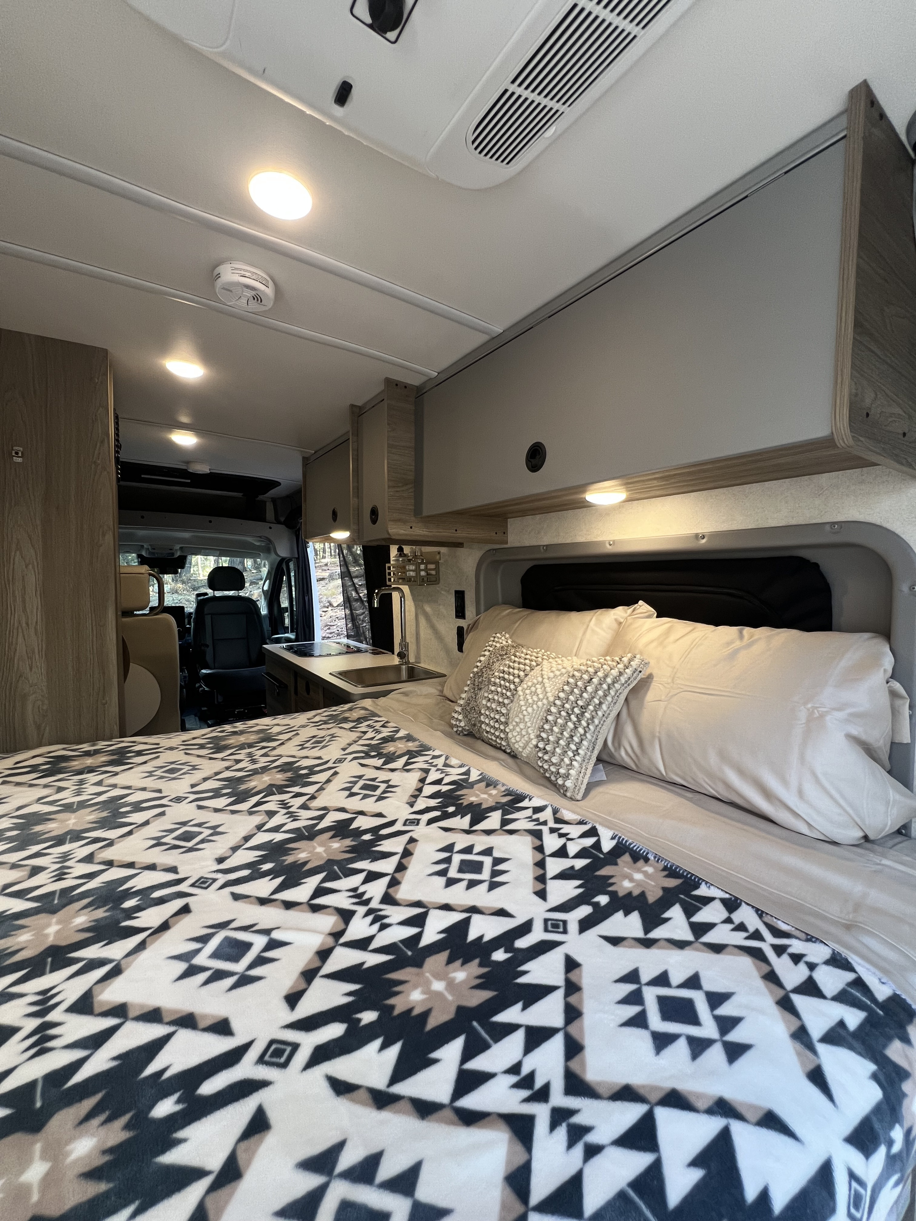 RV rental interior: bed with patterned blanket, compact kitchen, & overhead cabinets. Ready for travel!. Winnebago Solis 59PX 2025
