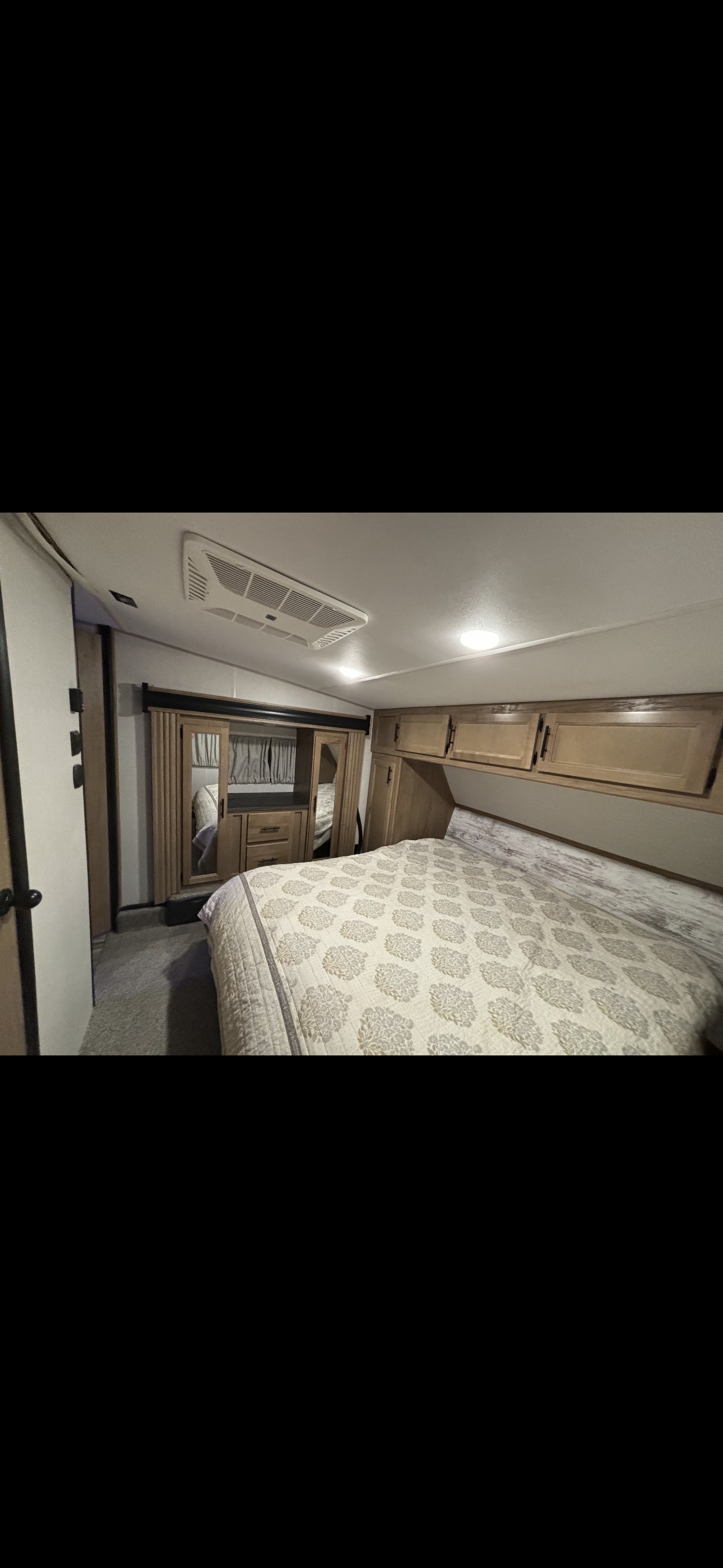 Inside an RV rental: a cozy bedroom features a patterned quilt on the bed, mirrored wardrobe, and overhead wooden cabinets.. Forest River 365 wolf pack toy hauler 2024