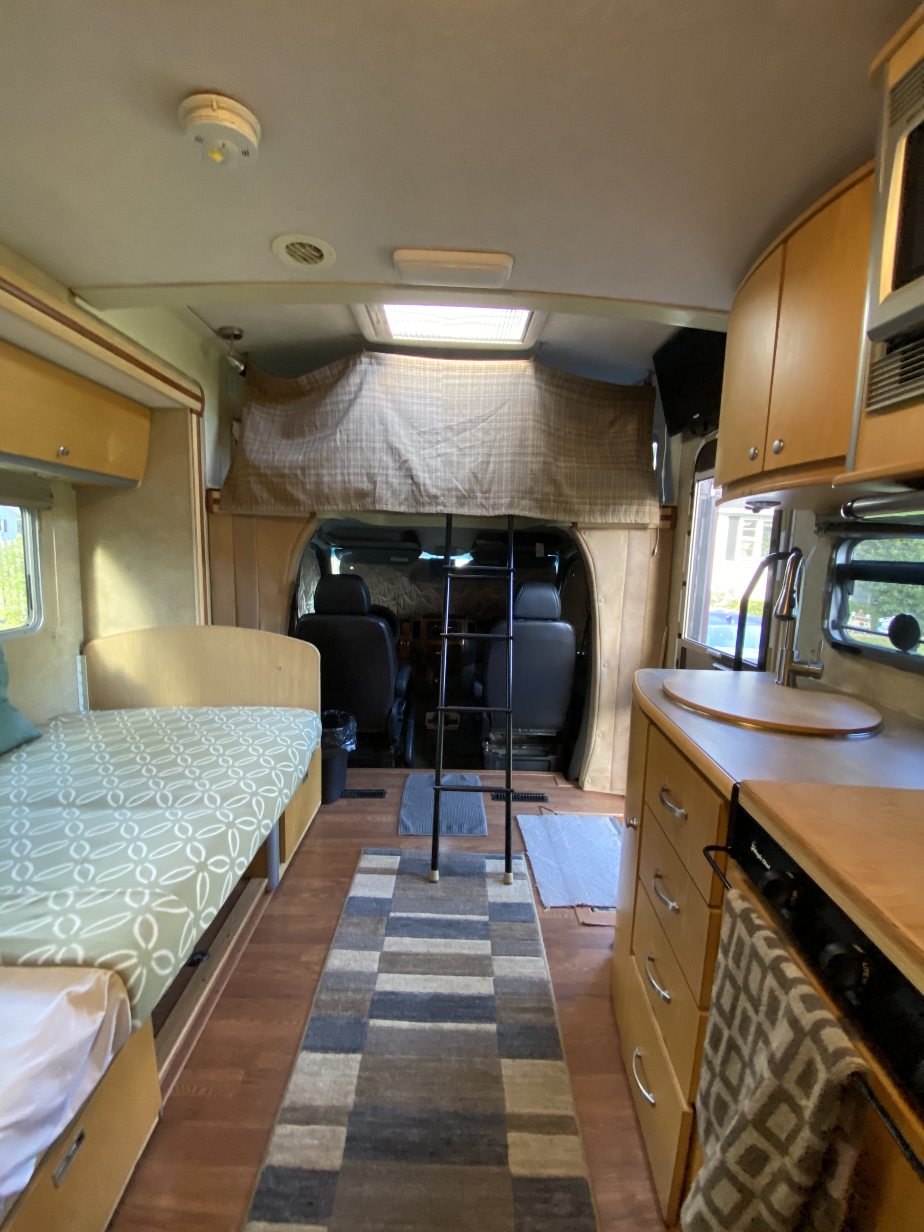 Inside an rv rental: a cozy bed, kitchenette with sink & stove, and a ladder leading to the sleeping loft above the cab.. Winnebago View 2008