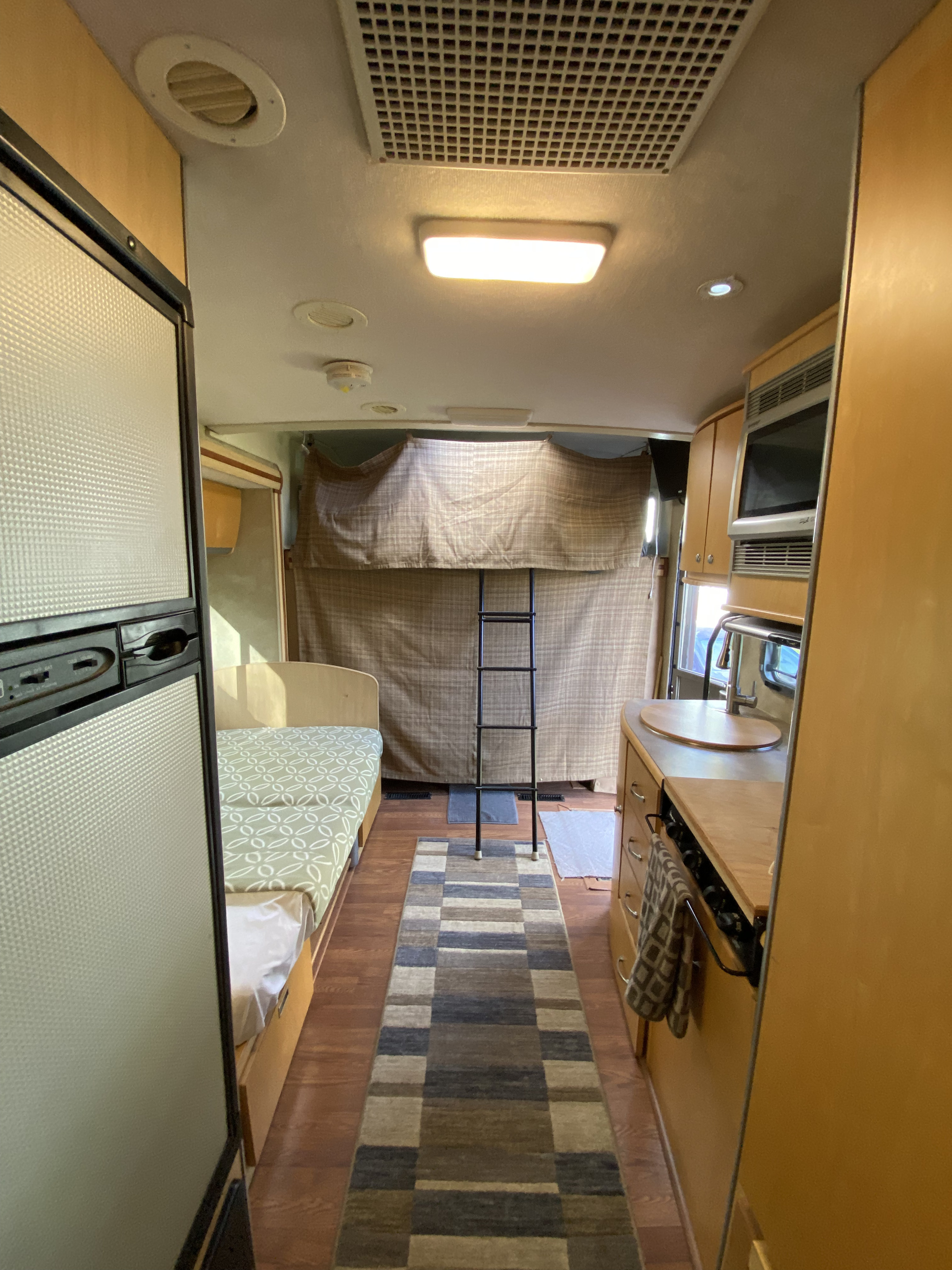 RV rental interior featuring a bed, kitchen with a microwave, bunk ladder, and a long patterned rug down the aisle.. Winnebago View 2008