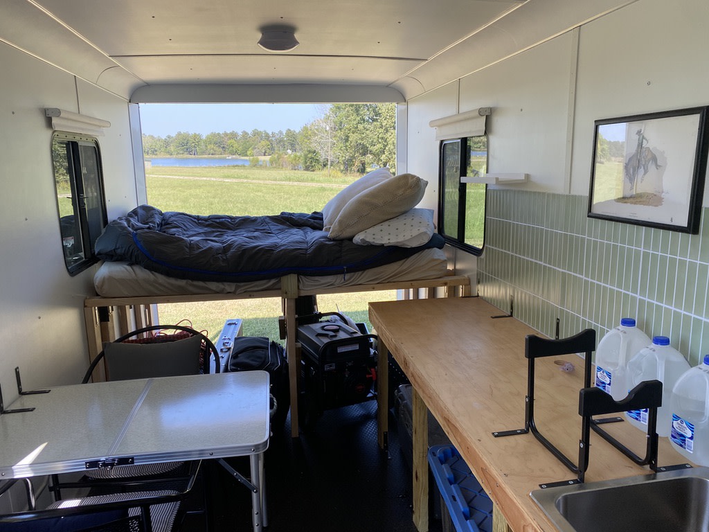 RV rental interior: elevated bed, kitchen counter with sink, and folding table, showcasing a scenic lake view.. Haulmark Transport V-Nose 2024