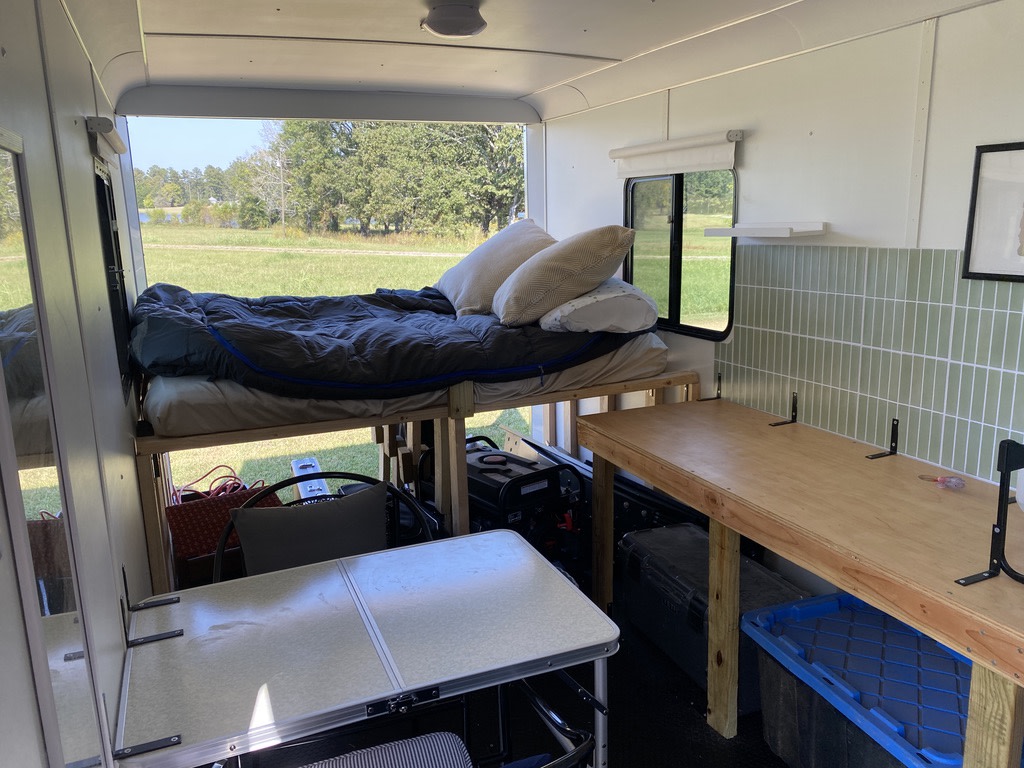 Cozy rv rental interior: raised bed, two tables, and storage solutions against a tiled wall, perfect for mobile living.. Haulmark Transport V-Nose 2024