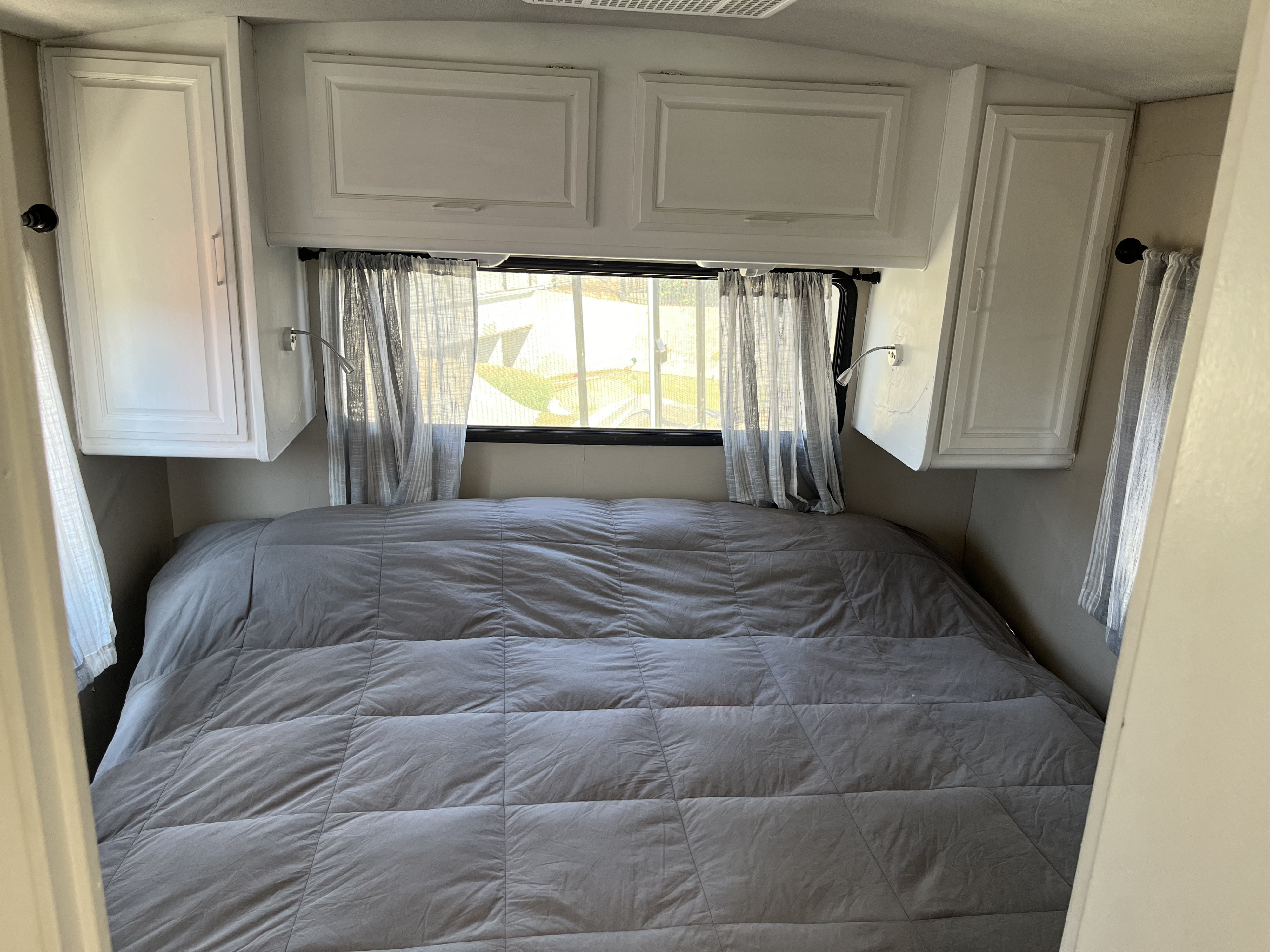 Cozy RV rental bedroom featuring a large grey quilted bed, white overhead cabinets, and sheer window curtains.. Fleetwood Bounder 1995