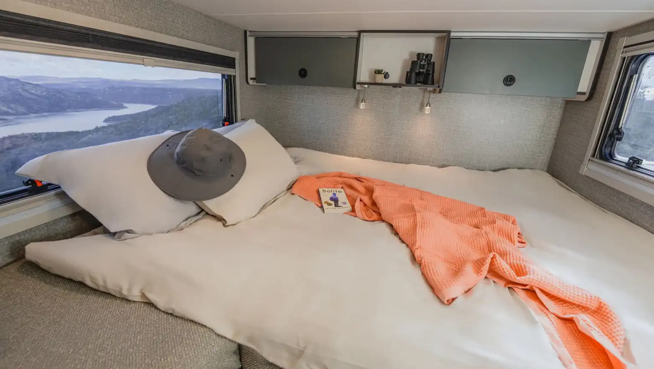 RV rental bedroom features a cozy bed with a hat & throw. A wide window offers stunning mountain and lake views.. Winnebago Ekko 2025