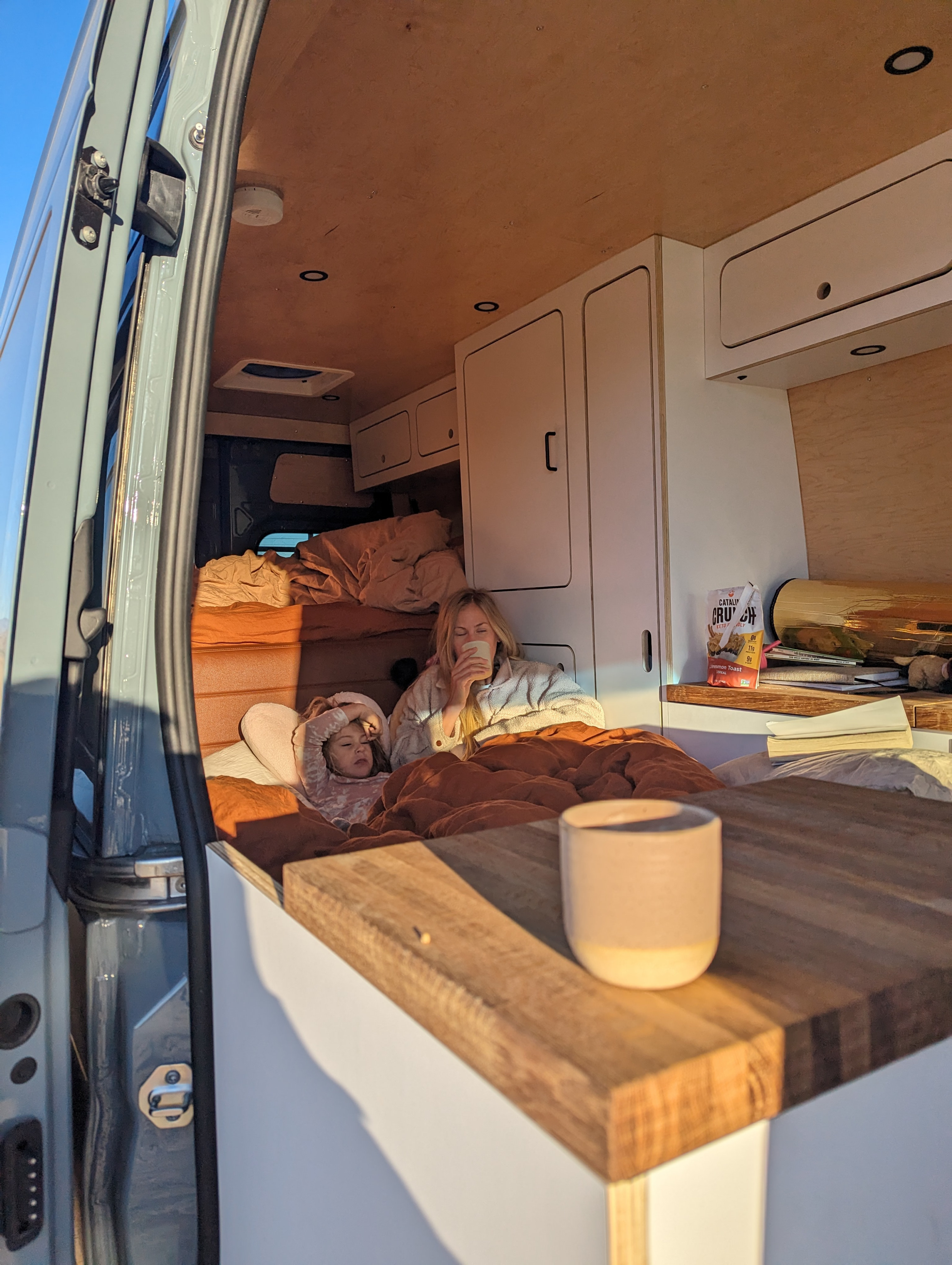 Cozy morning in an RV rental: a woman sips coffee while a child rests in bed inside a custom camper van. Perfect for family adventures.. Mercedes-Benz Sprinter 419 LWB High Roof 2023