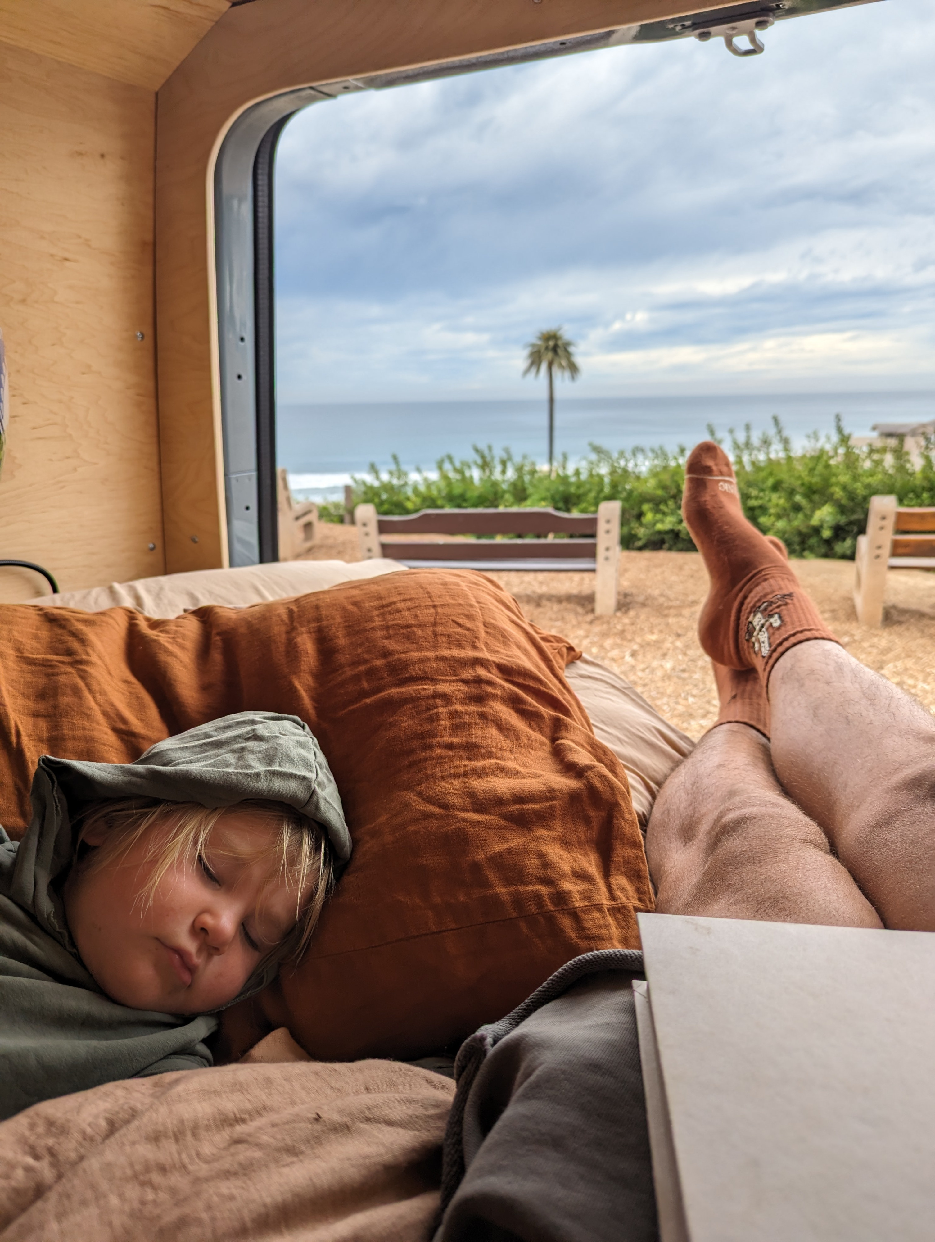 Relaxing in an RV rental: a child sleeps soundly while an adult's feet rest, overlooking a coastal ocean view.. Mercedes-Benz Sprinter 419 LWB High Roof 2023