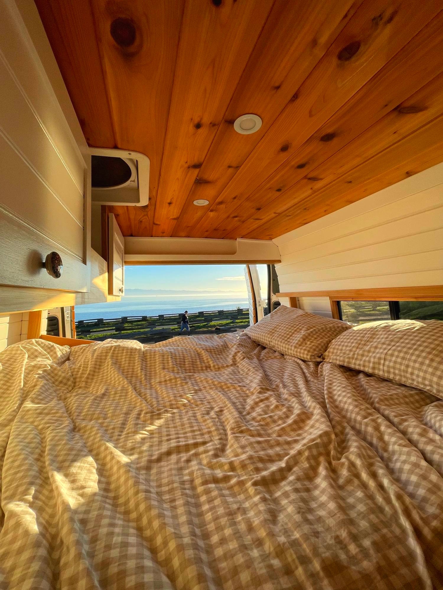 Cozy RV rental interior with a comfy bed and checkered duvet. Open back doors reveal a stunning ocean view.. Ram Promaster 3500 2018