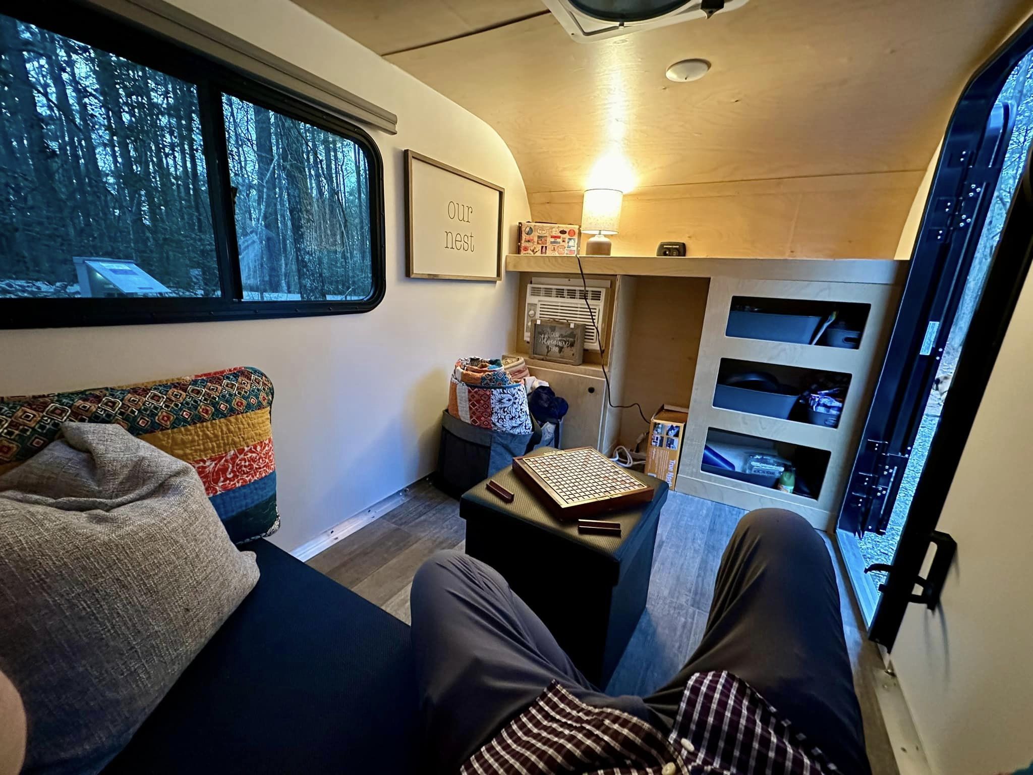 Serene RV rental interior: person relaxing on sofa, forest view, board game on ottoman, "Our Nest" sign.. Rustic Trails Koala 2023