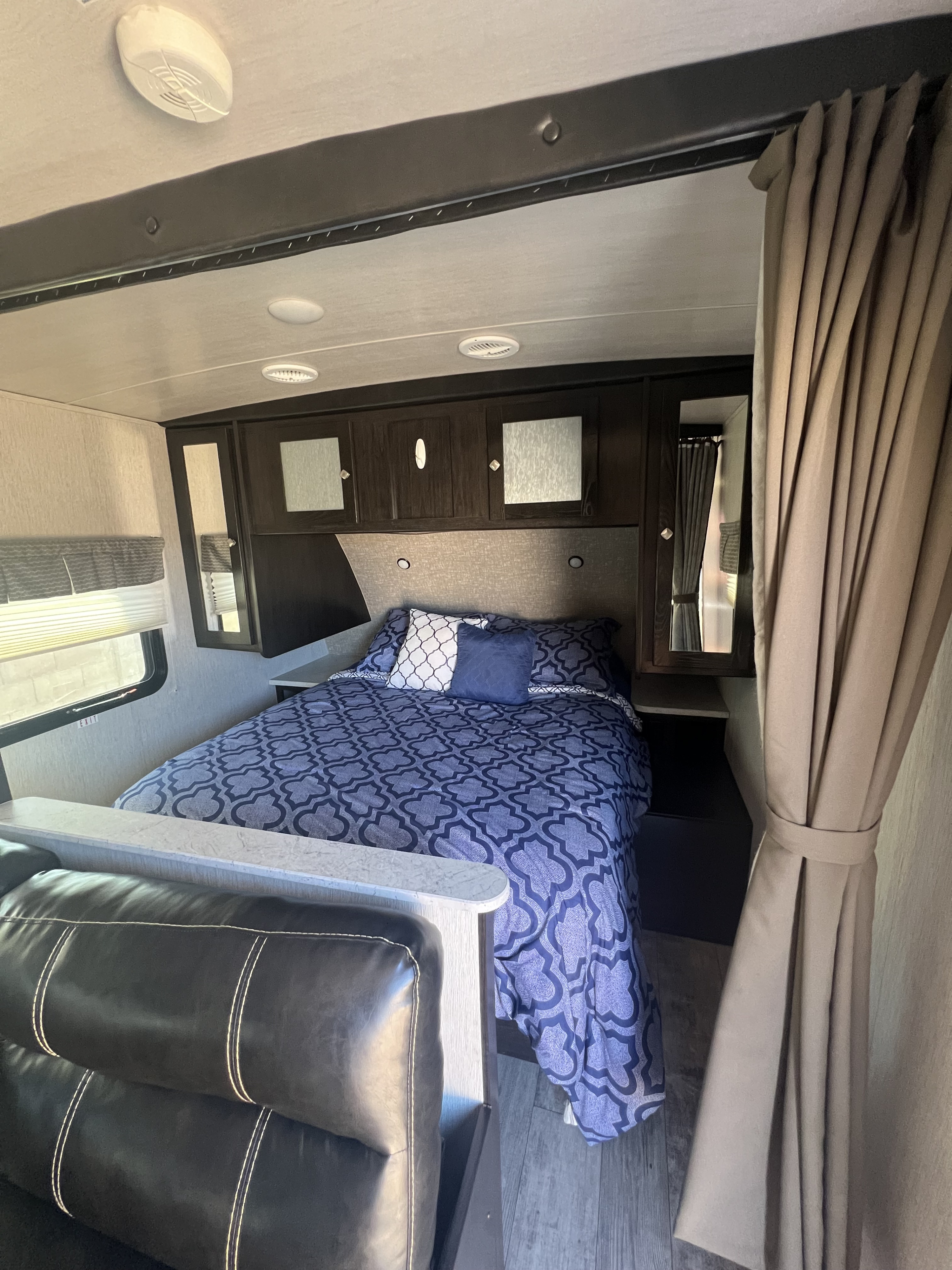 Cozy RV rental bedroom with patterned blue bedding, overhead cabinets, and adjacent black leather sofa.. Heartland RVs North Trail 2018