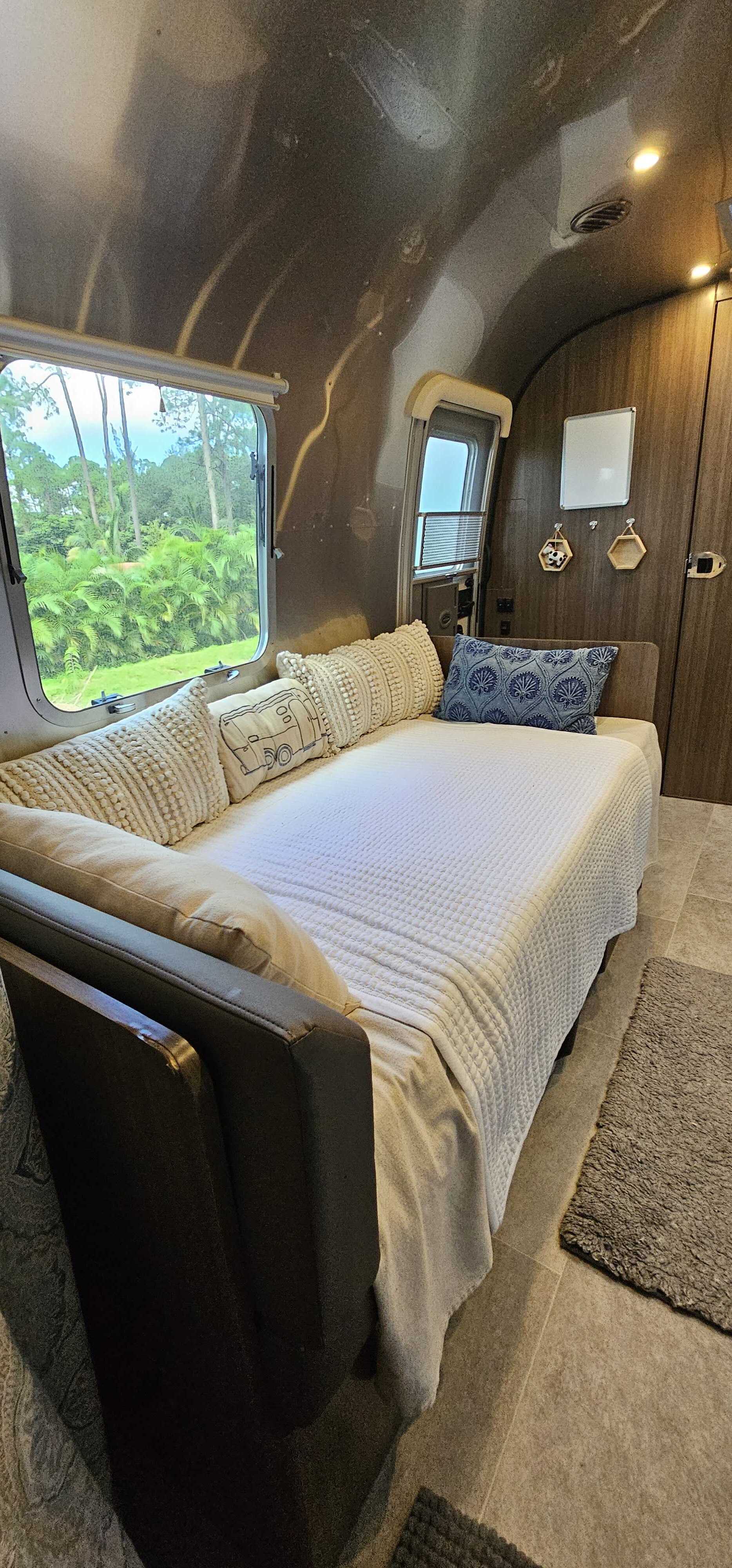 Step into a comfortable Airstream RV rental. Features a cozy daybed, decorative pillows, and large window with lush outdoor views.. Airstream Caravel 2022