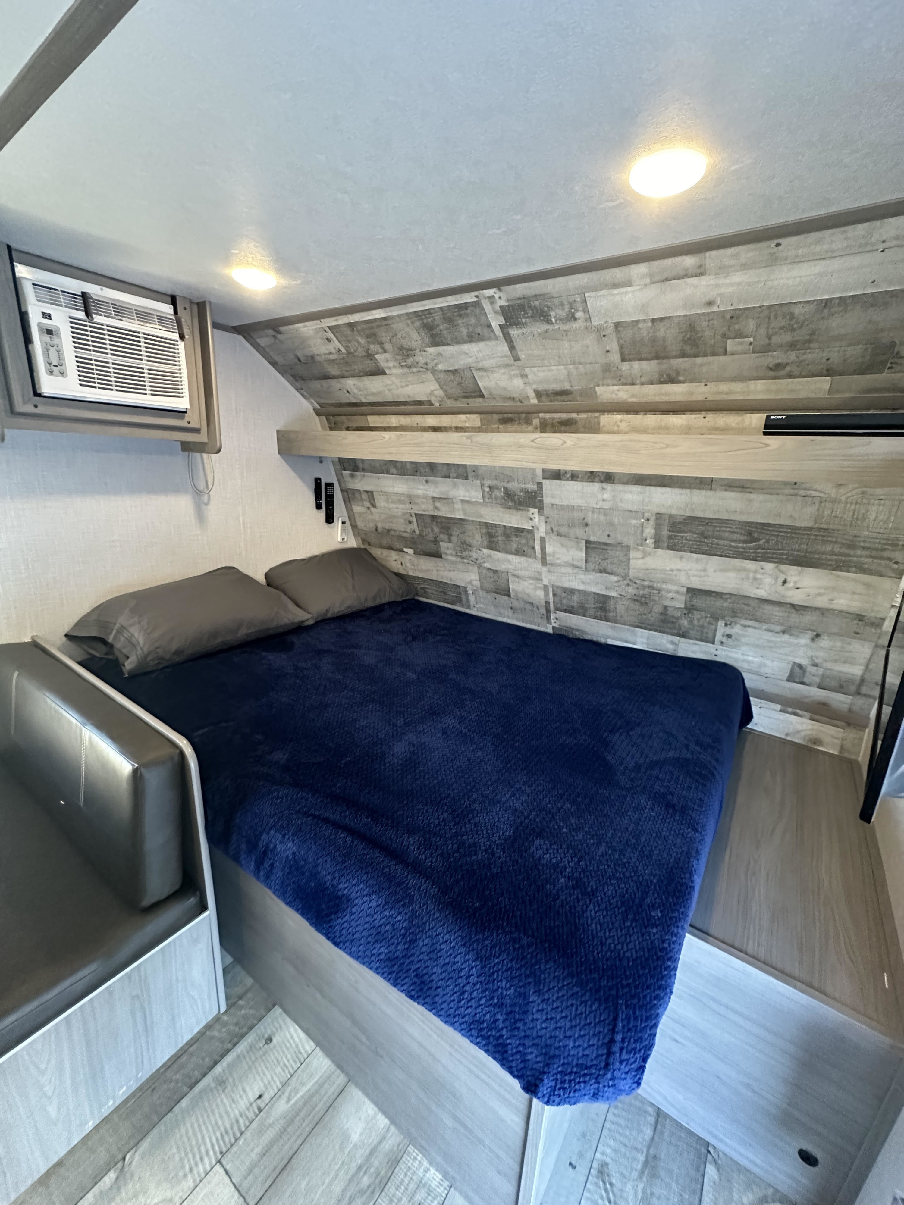 Cozy RV rental interior: bedroom with a blue-blanketed bed, mounted AC, and modern wood-look walls.. Dutchmen Coleman Lantern LT 2023