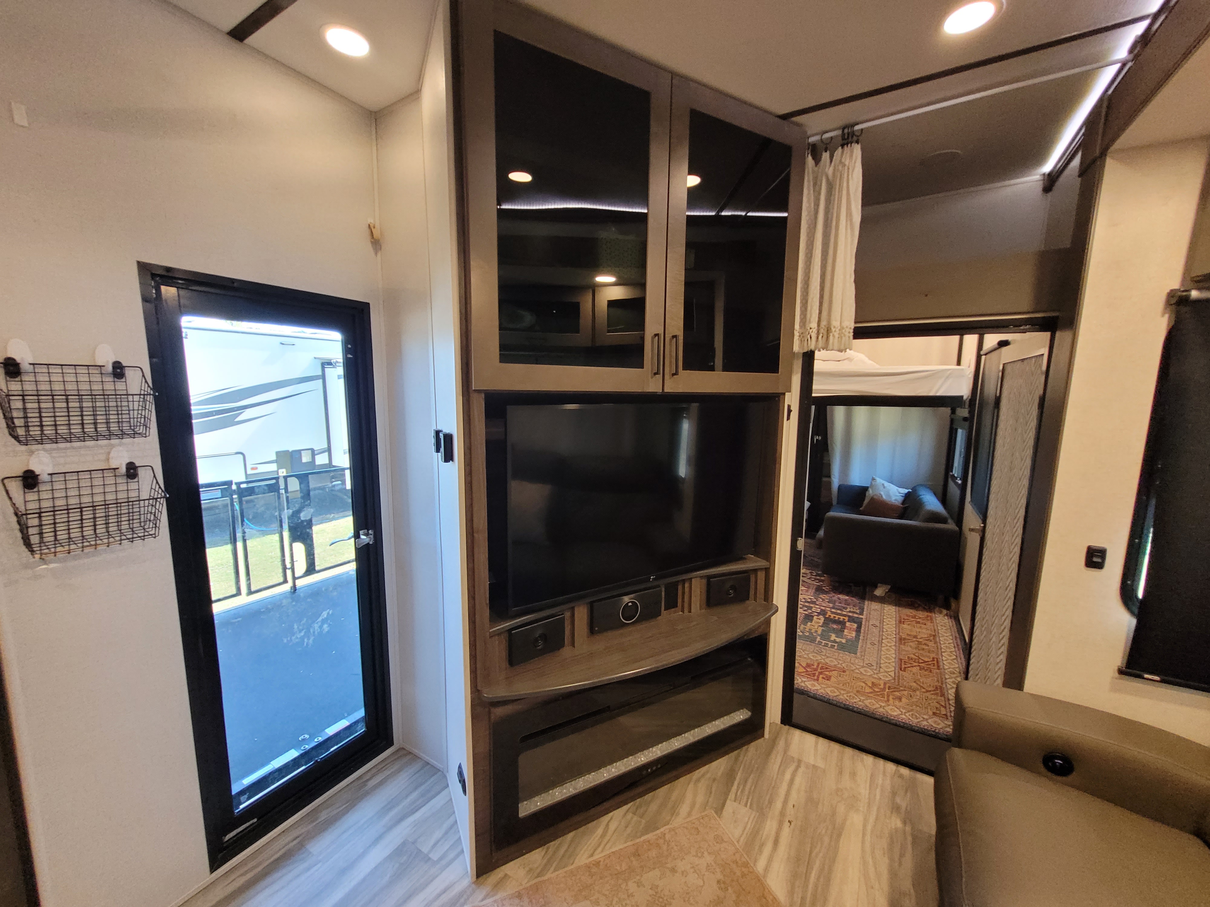 Modern RV rental interior: living area with TV, fireplace, glass-door cabinets, and wall baskets. Entry to bunk room and outdoor patio.. Alliance Valor 2023