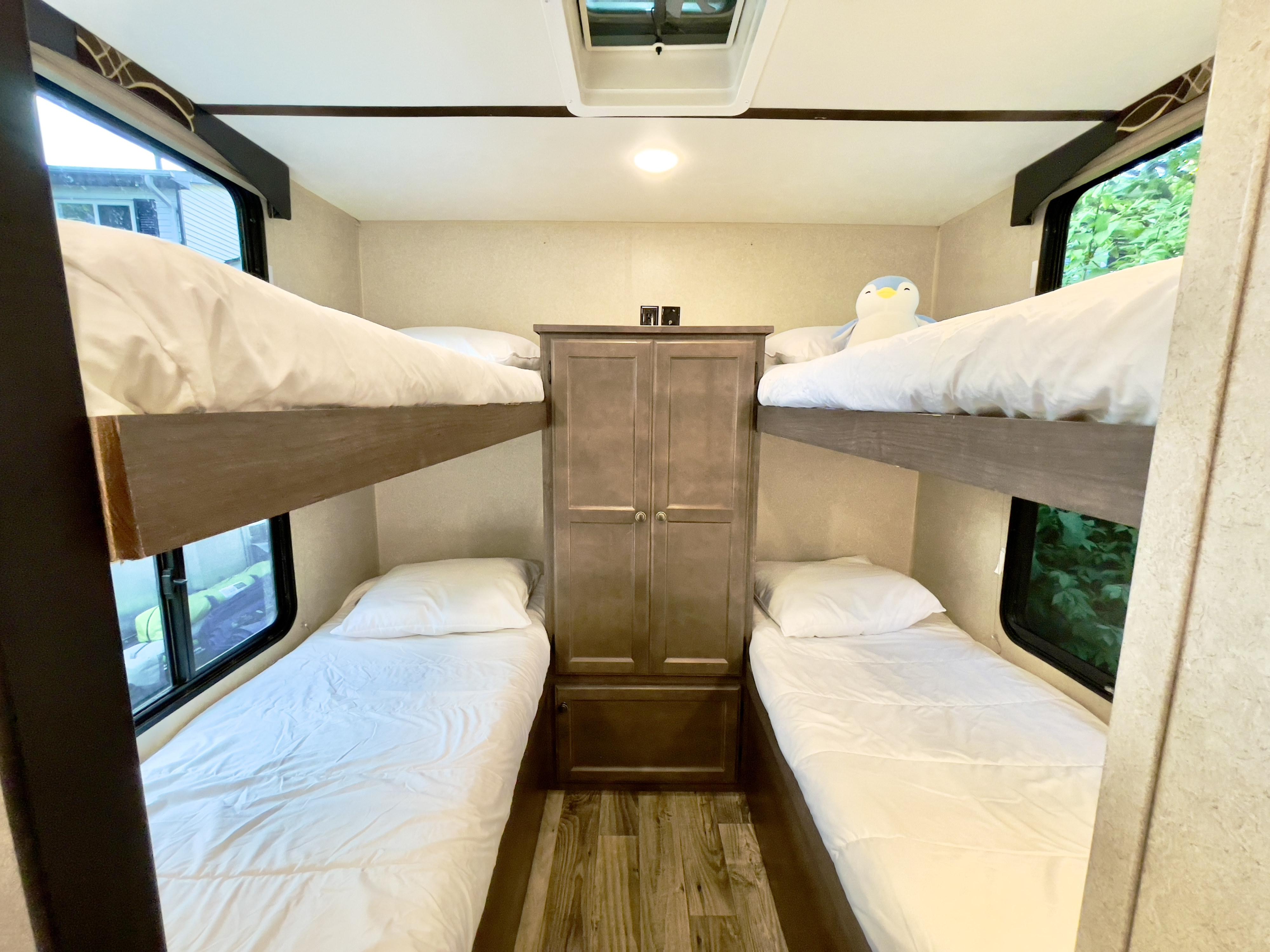 Interior of an RV rental: four bunk beds with white bedding, a central wardrobe, and windows looking out.. Sunset Park RV Sun Lite 2019