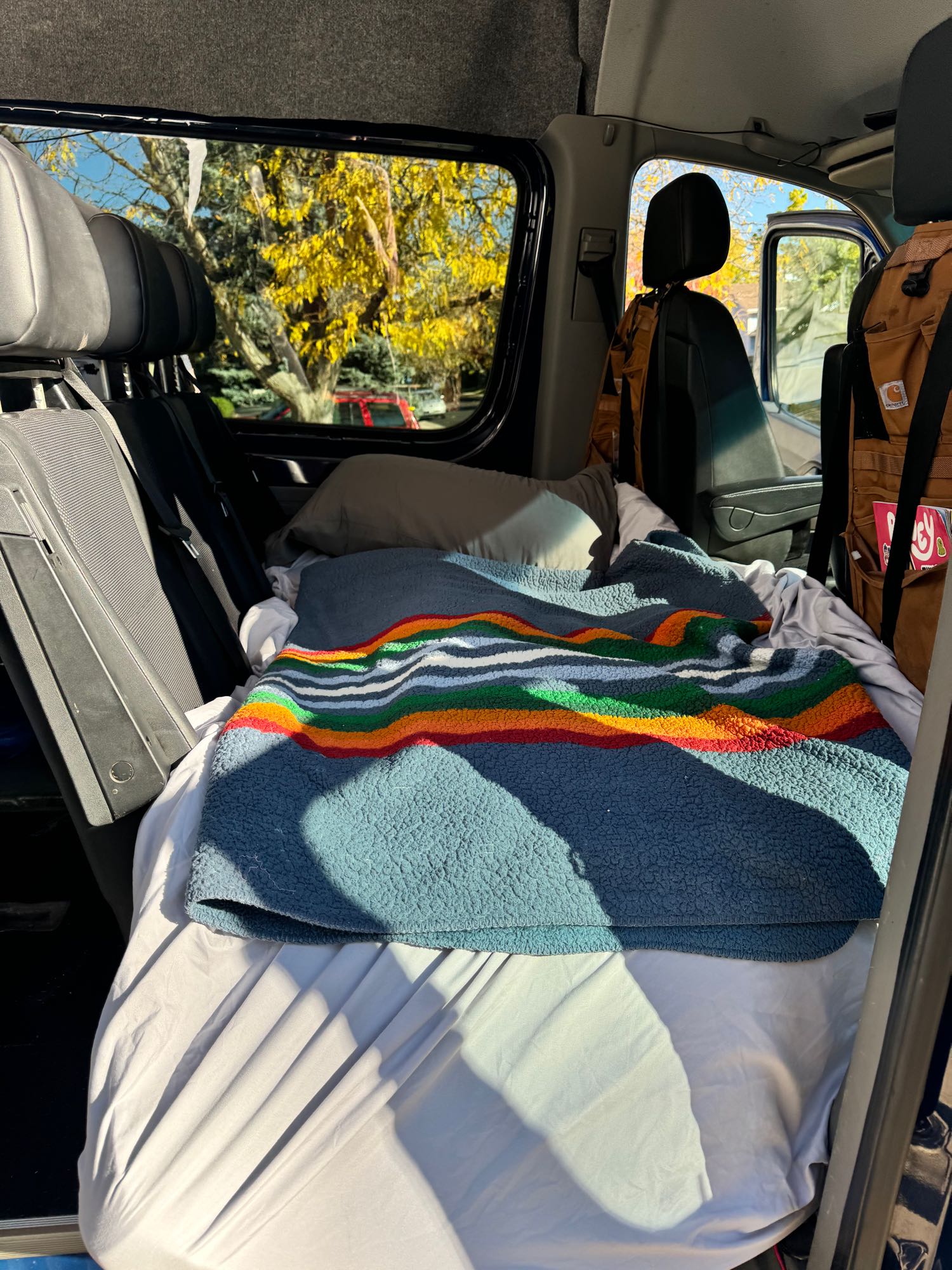 Cozy bed inside an RV rental, covered with a colorful striped blanket. Autumn trees visible through sunlit windows.. Mercedes-Benz Sprinter 2016