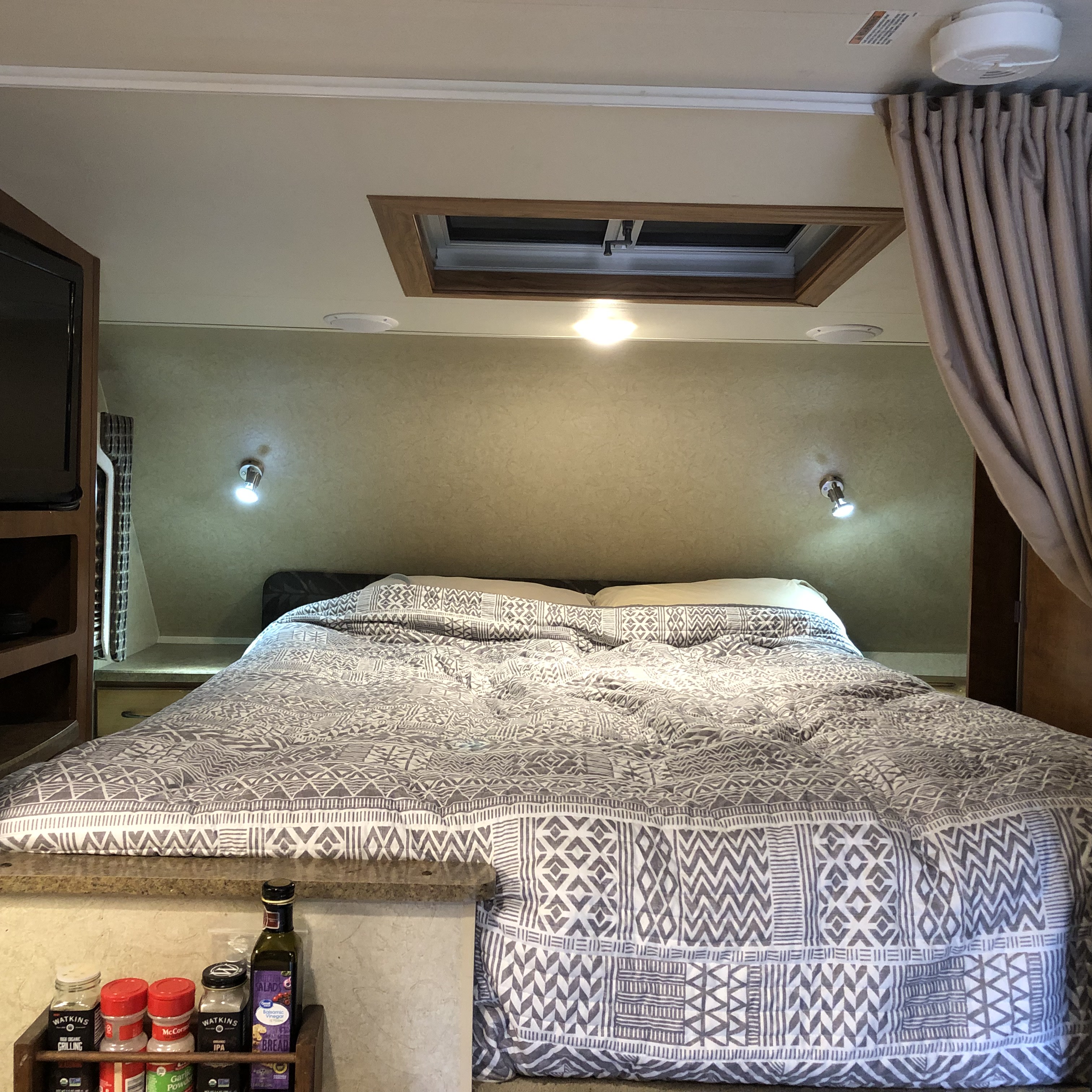 Inside an RV rental: a comfortable bed with patterned bedding, overhead skylight, TV, and spices nearby.. Lance 855s 2016