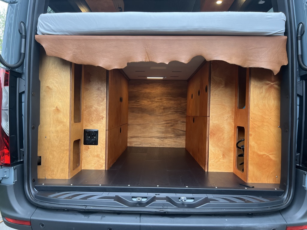 Rear view of an RV rental's custom interior: raised bed with leather trim, wood storage, and dark floor for adventures.. Mercedes Sprinter 2500 Turbo Diesel 2023