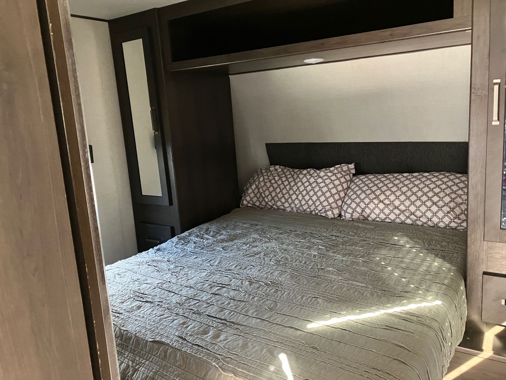 RV rental bedroom featuring a comfortable bed with patterned pillows, overhead storage, and a mirror wardrobe. Sunlight fills the space.. Grand Design Momentum Toy Hauler 2024