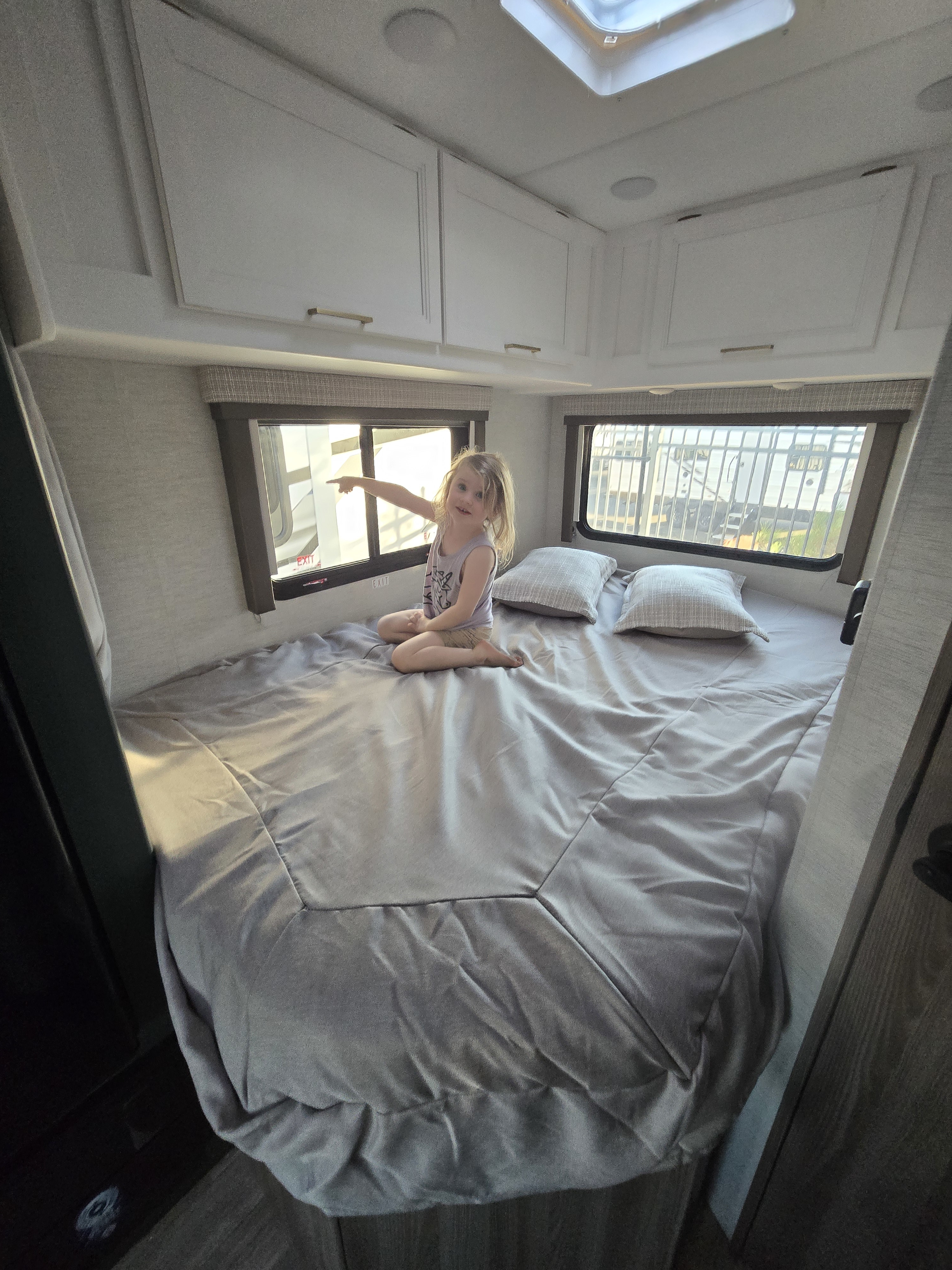 Cozy RV rental bedroom with a child pointing out the window from the large bed. Overhead cabinets complete the travel setup.. Thor Eddie Bauer 22EB 2025