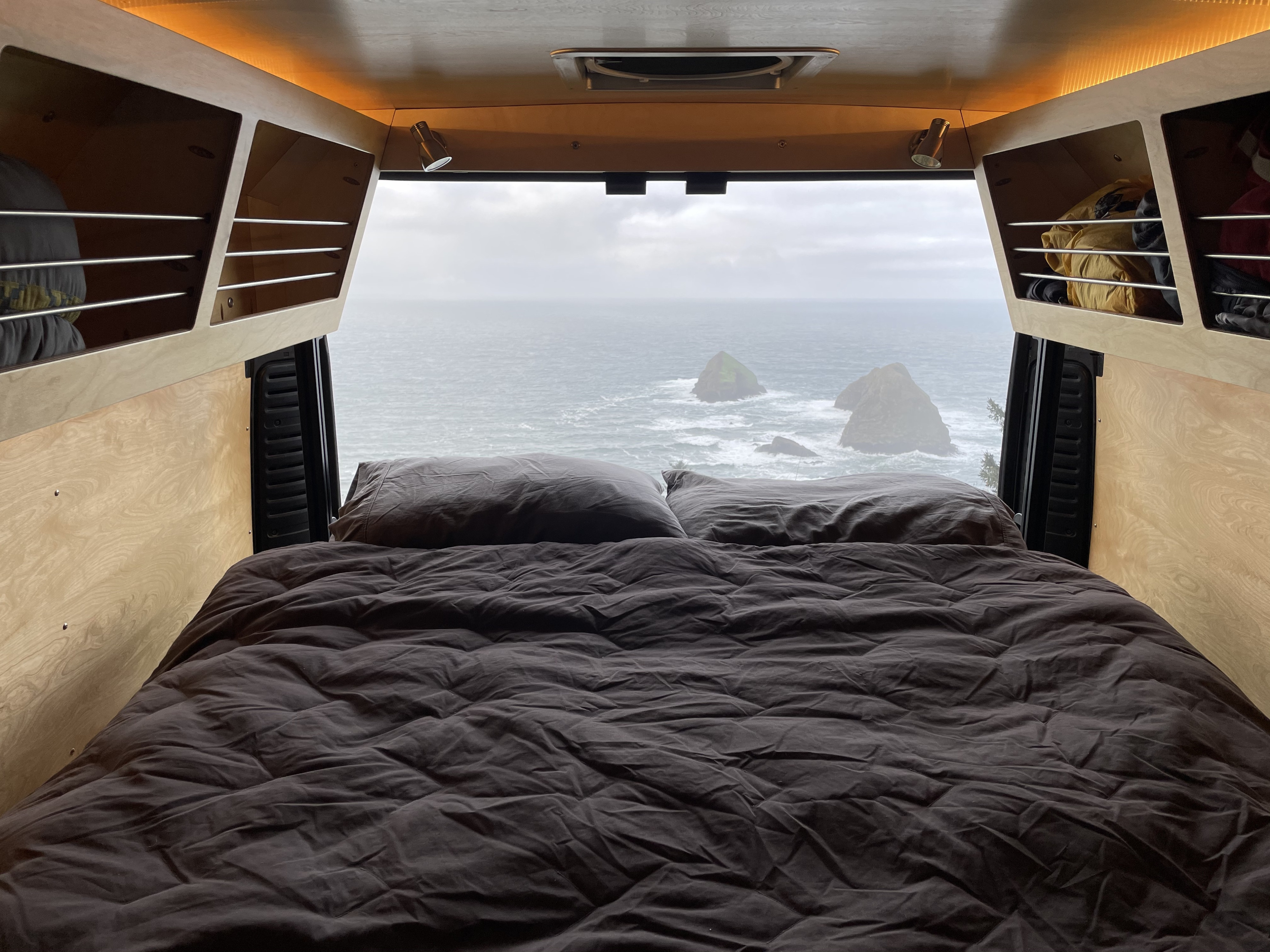 Inside a cozy RV rental, a bed faces a stunning ocean view with prominent sea stacks under a cloudy sky.. Ram Ram Promaster 2500 2018