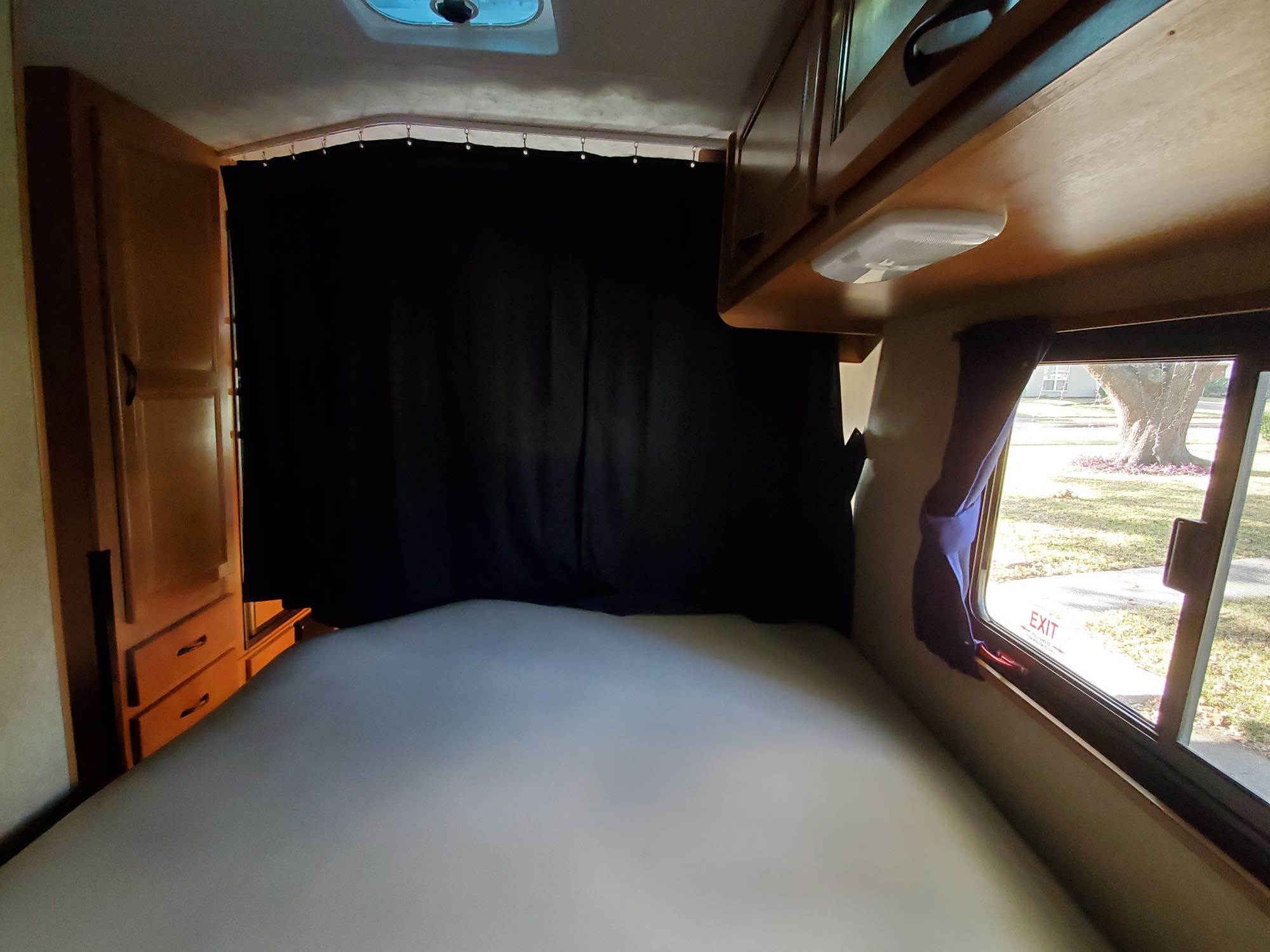 Inside a compact RV rental bedroom with a white mattress, dark privacy curtain, wooden cabinets, and window.. Thor Motor Coach Four Winds Majestic 2016
