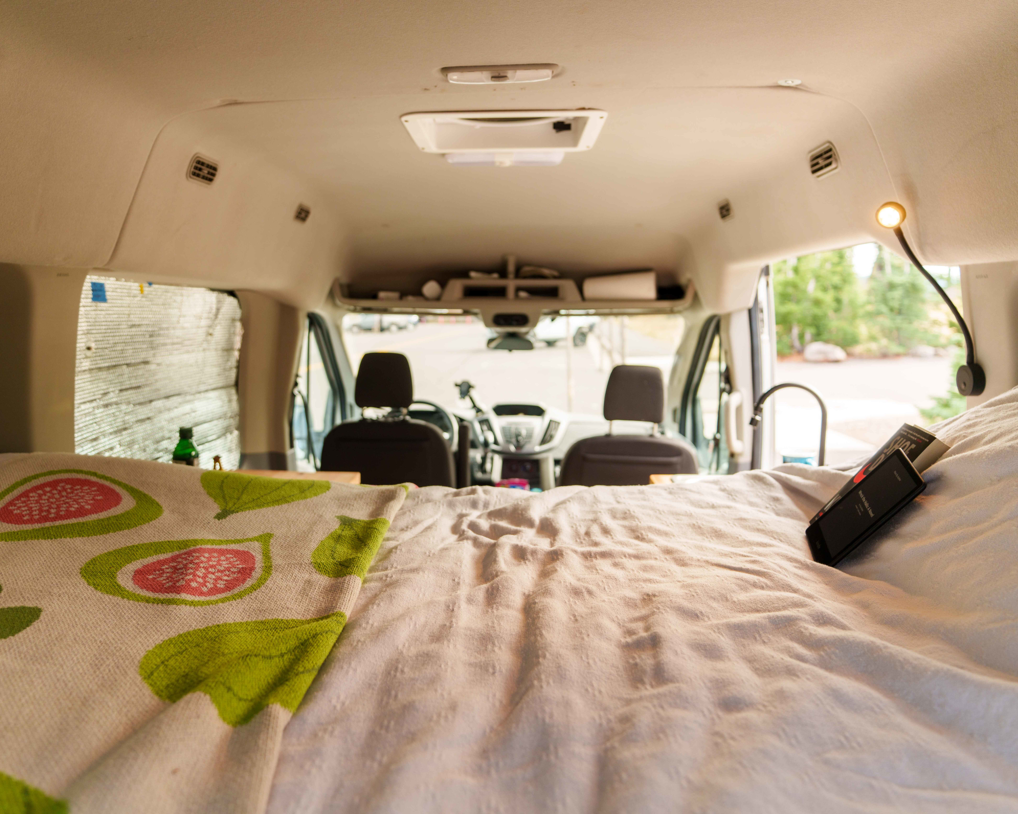 Inside a comfortable RV rental camper van looking forward. Bed in foreground with book/phone. Dashboard & outdoor view beyond.. Ford Transit 2018