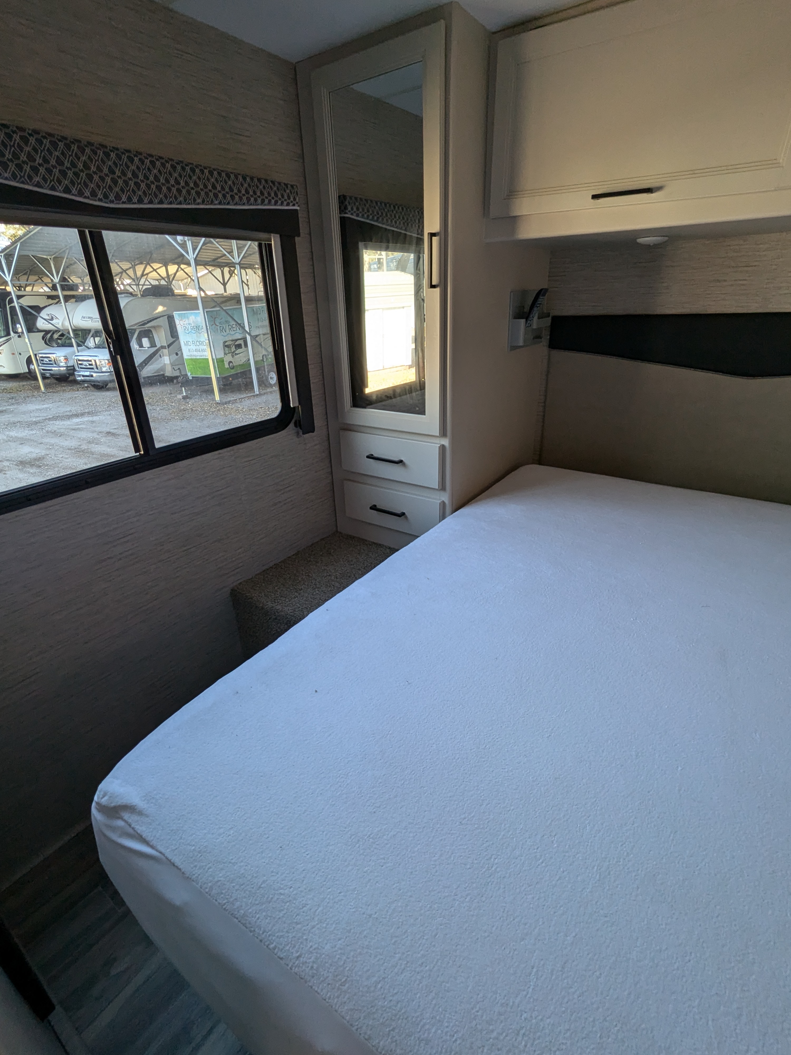 RV rental bedroom interior with a white bed, built-in mirror & cabinets, and a window view of parked RVs.. Thor Motor Coach Four Winds 2022