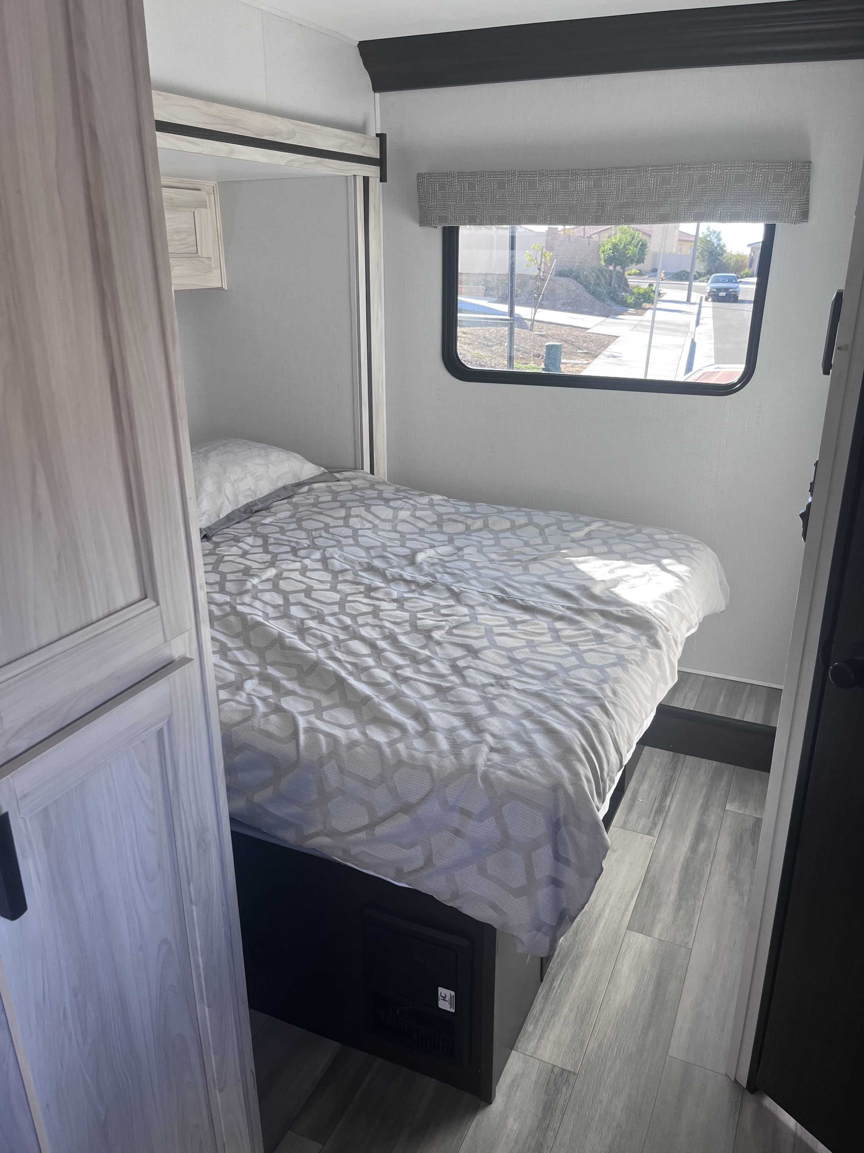 Cozy RV rental bedroom with gray patterned bedding, light wood-look interior, and a window view of a street.. Forest River Sunseeker LE 2023