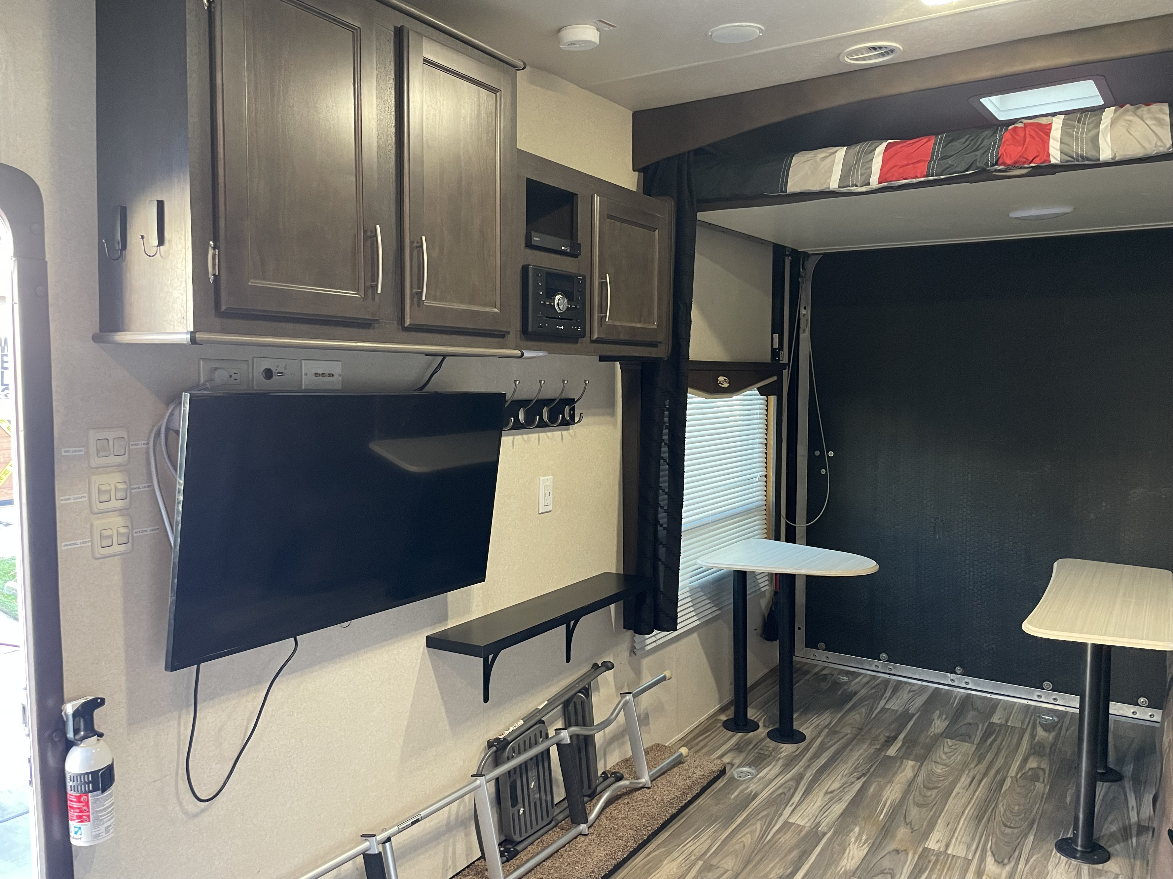 Inside a practical RV rental, featuring a TV, dark wood cabinets, overhead bunk with bedding, and two portable dining tables.. Forest River Stealth 2017