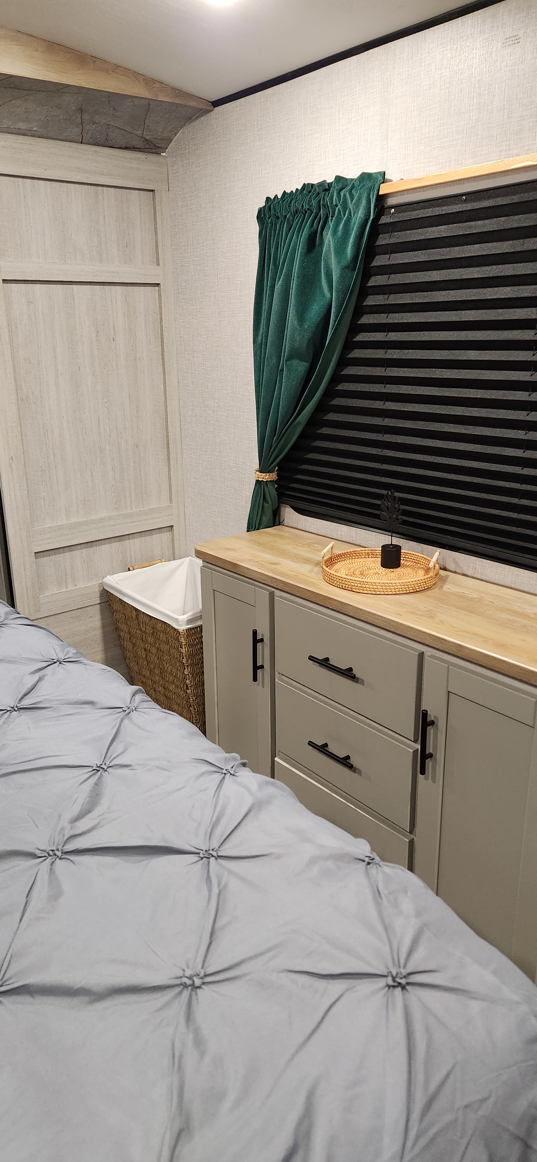 Inside an RV rental: a modern bedroom with a bed, dresser, wicker hamper, and window with dark green curtain and pleated blind.. Crossroads RV SS330SI 2024