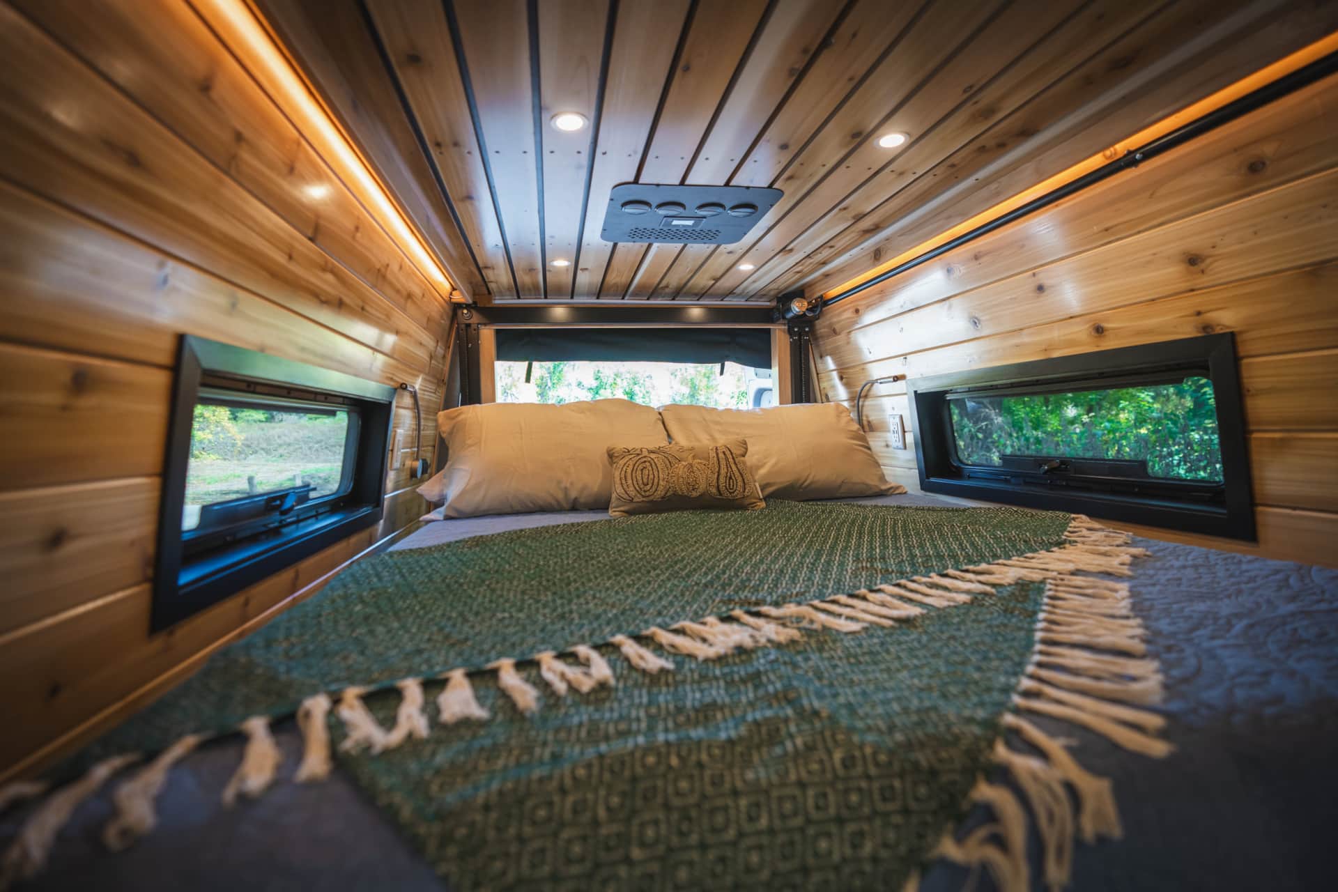Inside a comfortable RV rental bedroom, featuring warm wood walls, ceiling lights, and scenic windows.. Mercedes Sprinter 2023