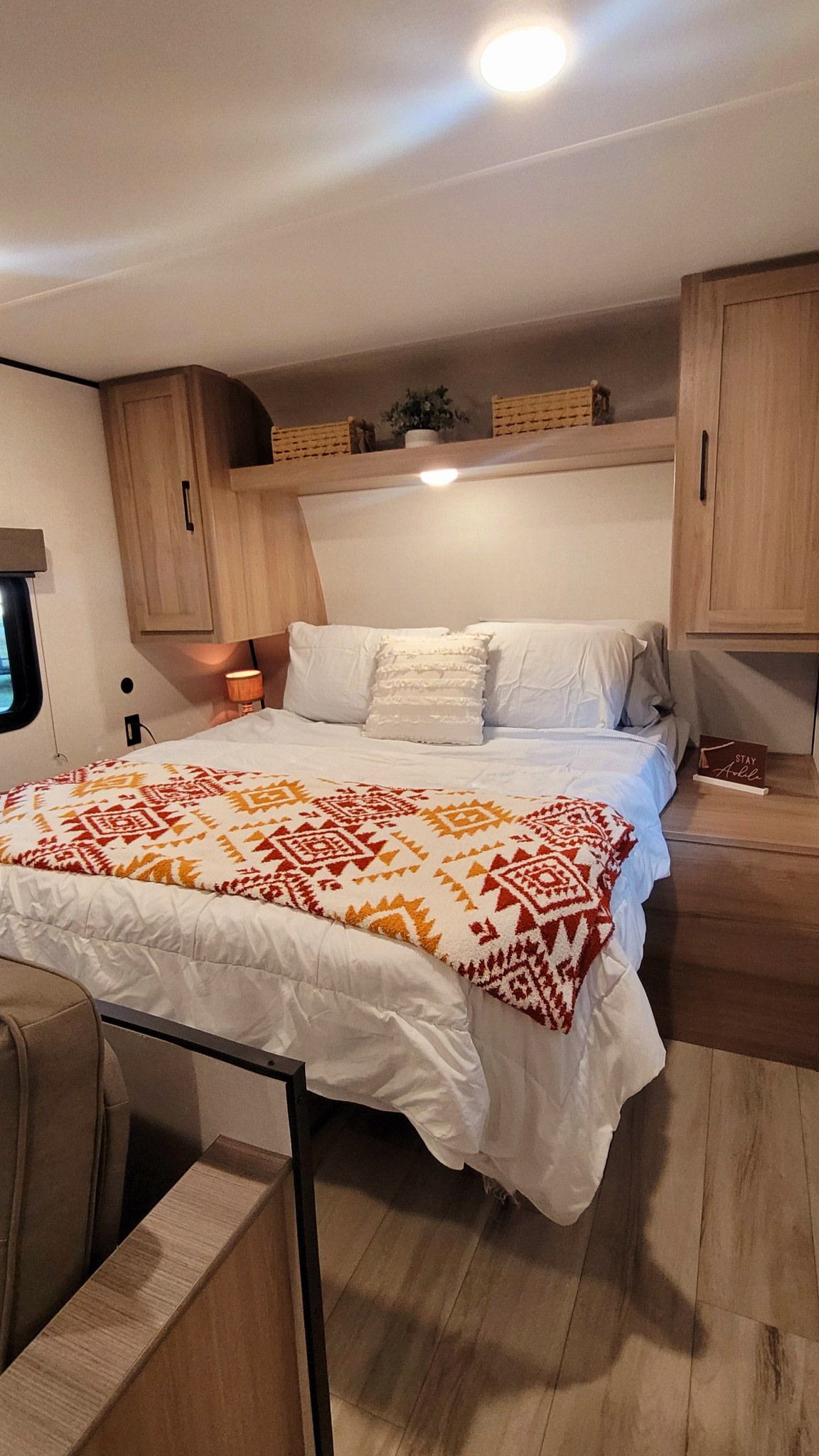 Cozy RV rental bedroom with a large bed, patterned blanket, overhead cabinets, and warm lighting.. keystone 23b 2025