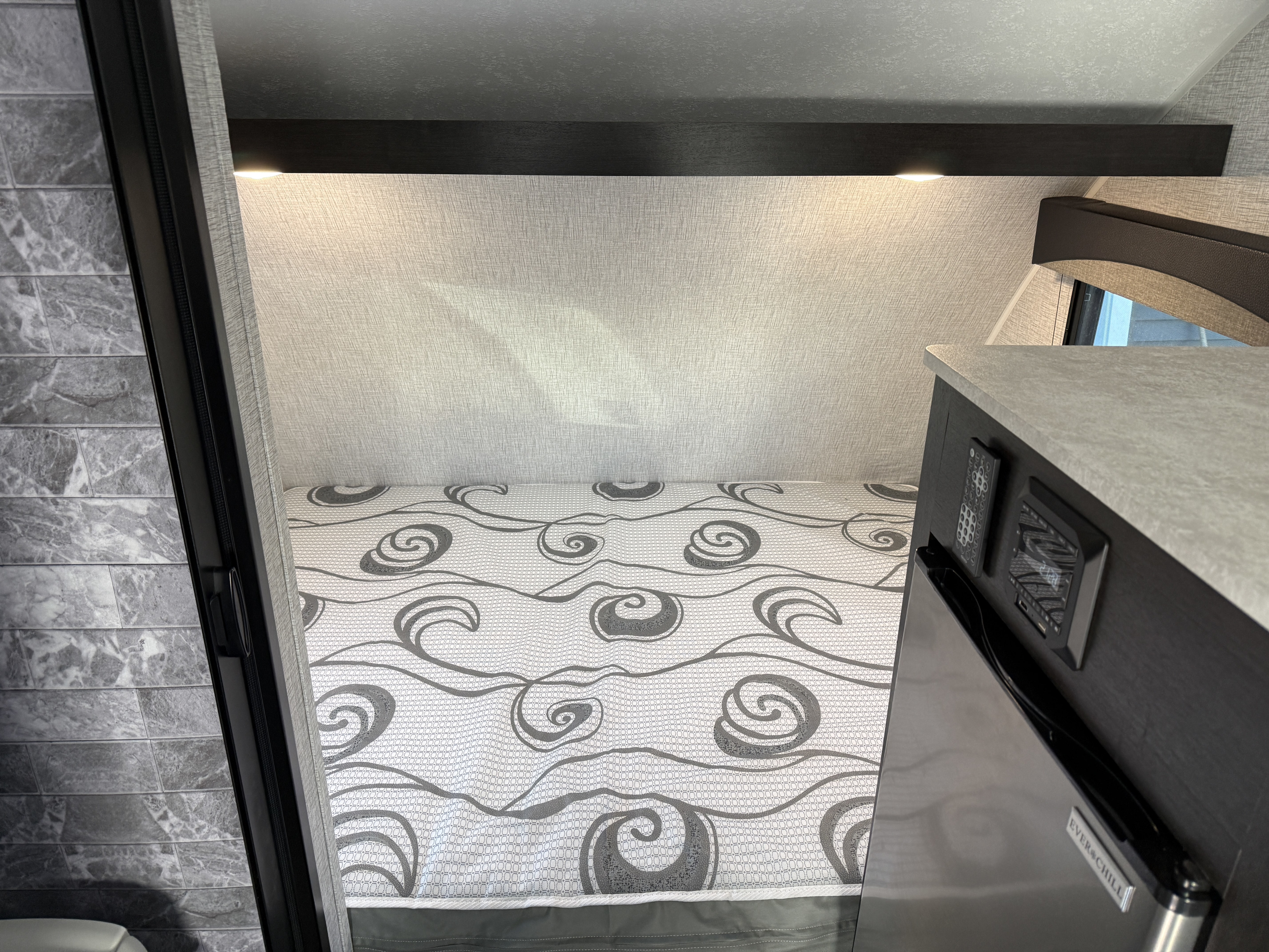 Modern RV rental interior: cozy sleeping area with a stylish patterned bed and compact fridge.. Braxton Creek Bushwhacker Plus 2022