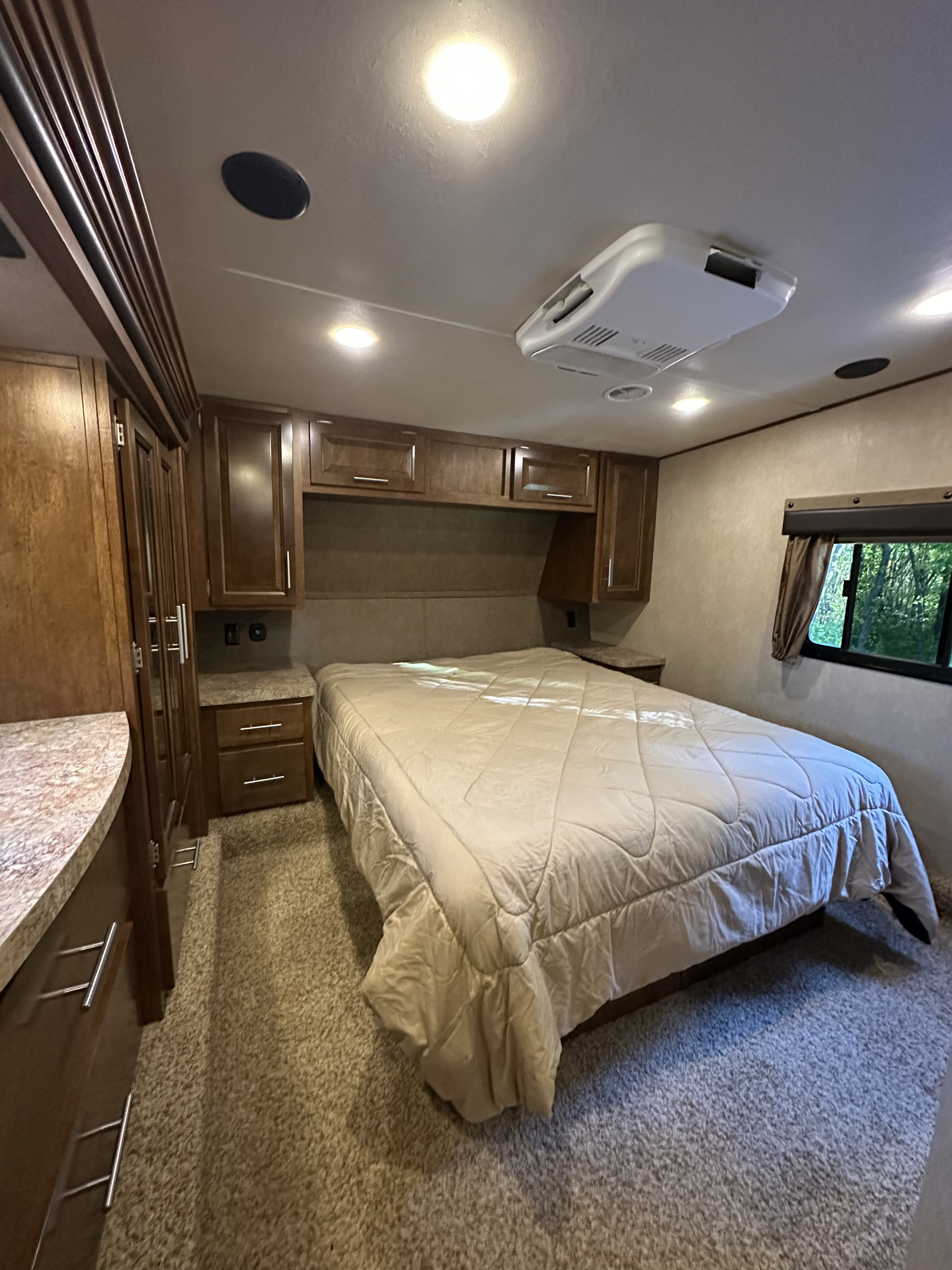 Inside an RV rental bedroom: cozy bed, wooden cabinets, window view of trees, AC unit & lights on ceiling.. Forest River Nitro Xlr 2015