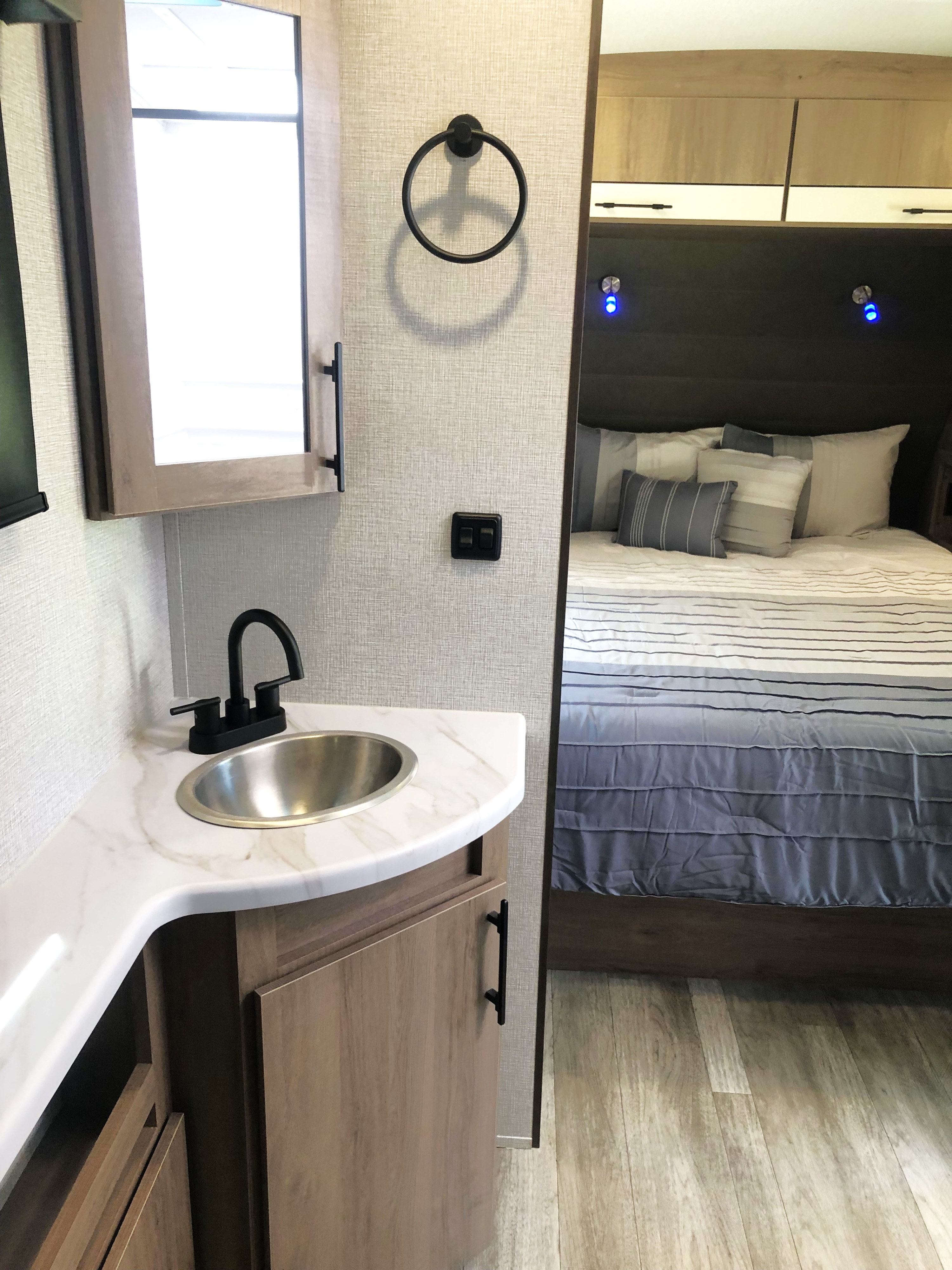 Compact RV rental interior featuring a modern bathroom vanity with sink and medicine cabinet, next to a cozy bedroom.. Grand Design Imagine 2023