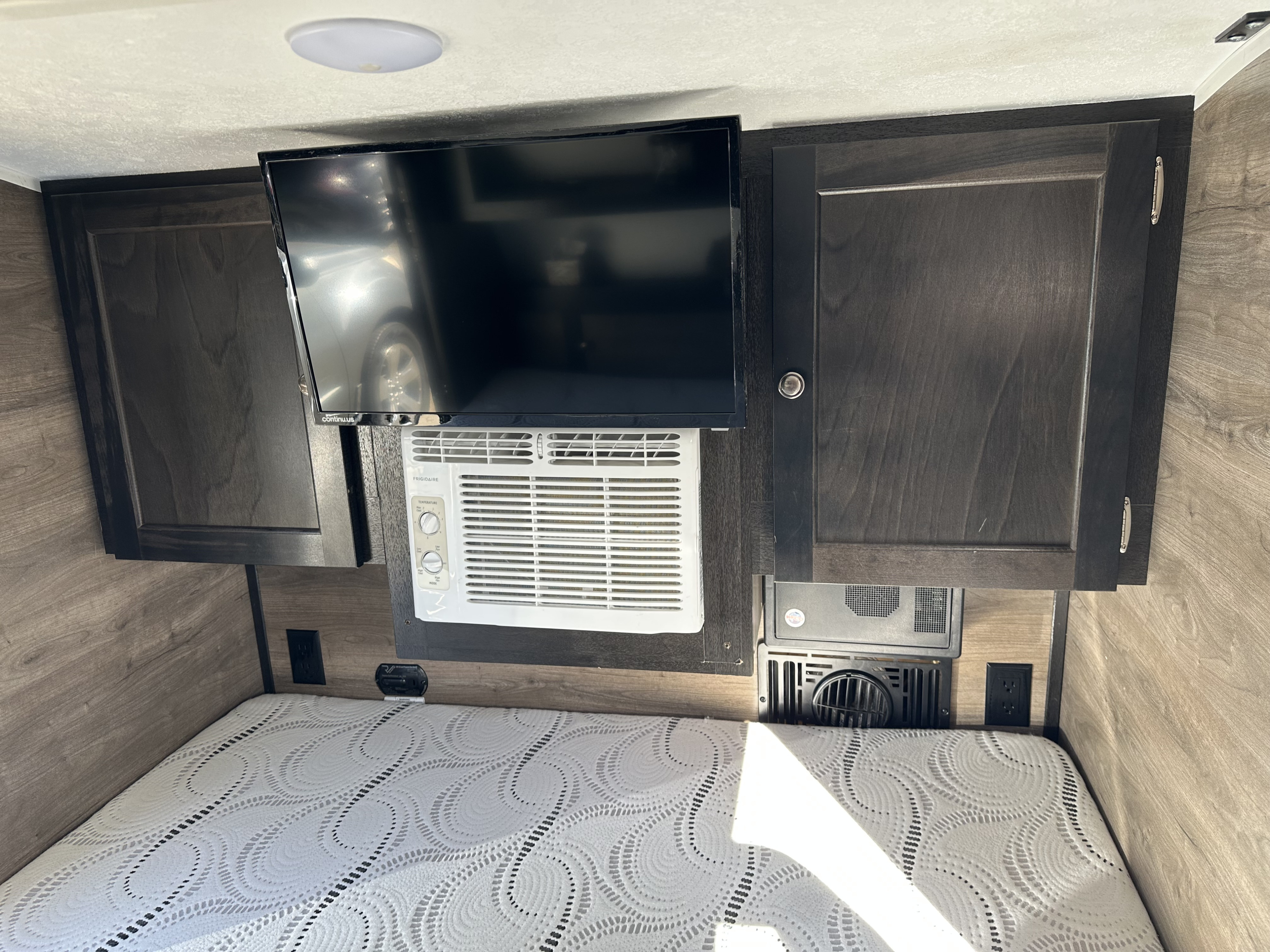 Inside an rv rental: Cozy sleeping area with a bed, mounted TV, Frigidaire AC unit, dark wood cabinets, and outlets.. Braxton Creek Bushwhacker 2021