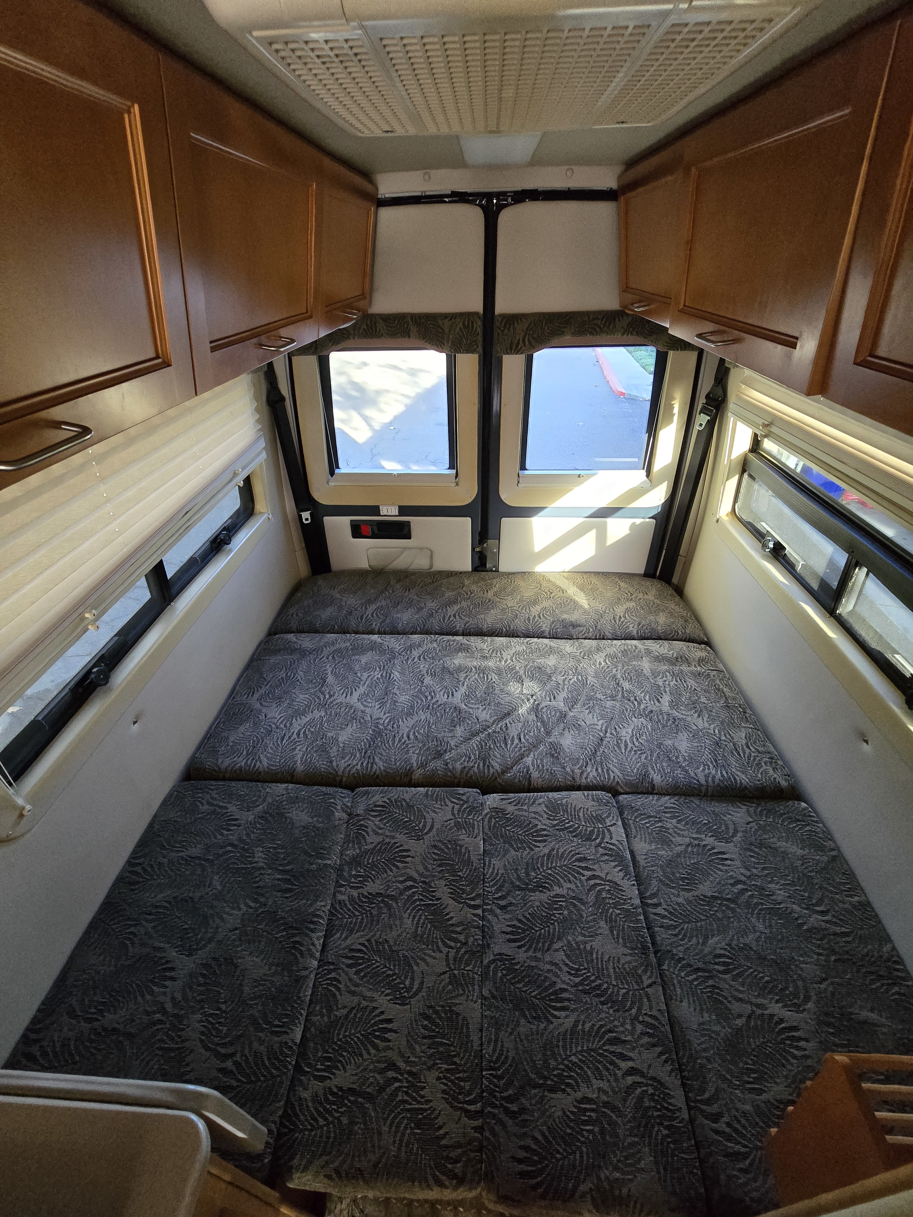 Inside an RV rental: a plush bed with leaf-patterned cushions, surrounding wood cabinets, and bright windows.. Leisure Travel Vans Leisure Travel Vans Class B 2004
