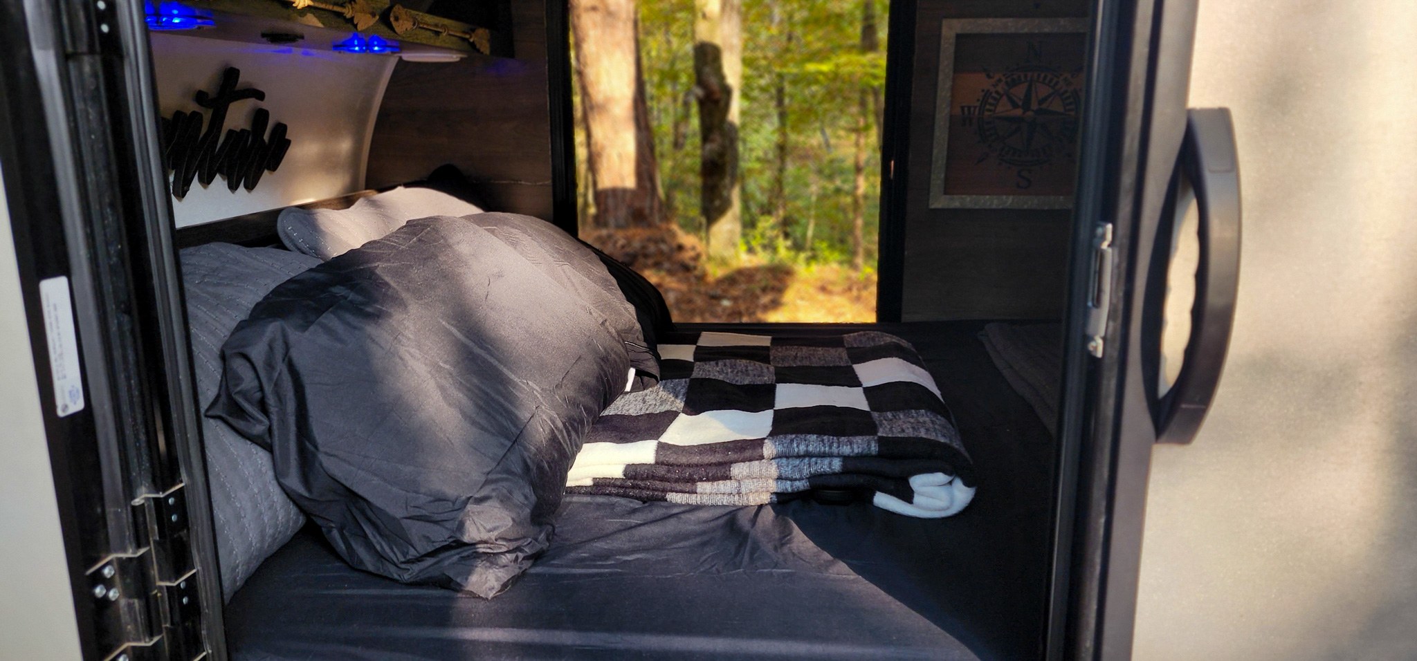 Step inside this cozy RV rental! Features a dark bed with a checkered blanket, blue lights, and a window view of the forest.. Braxton Creek Bushwhacker 2021