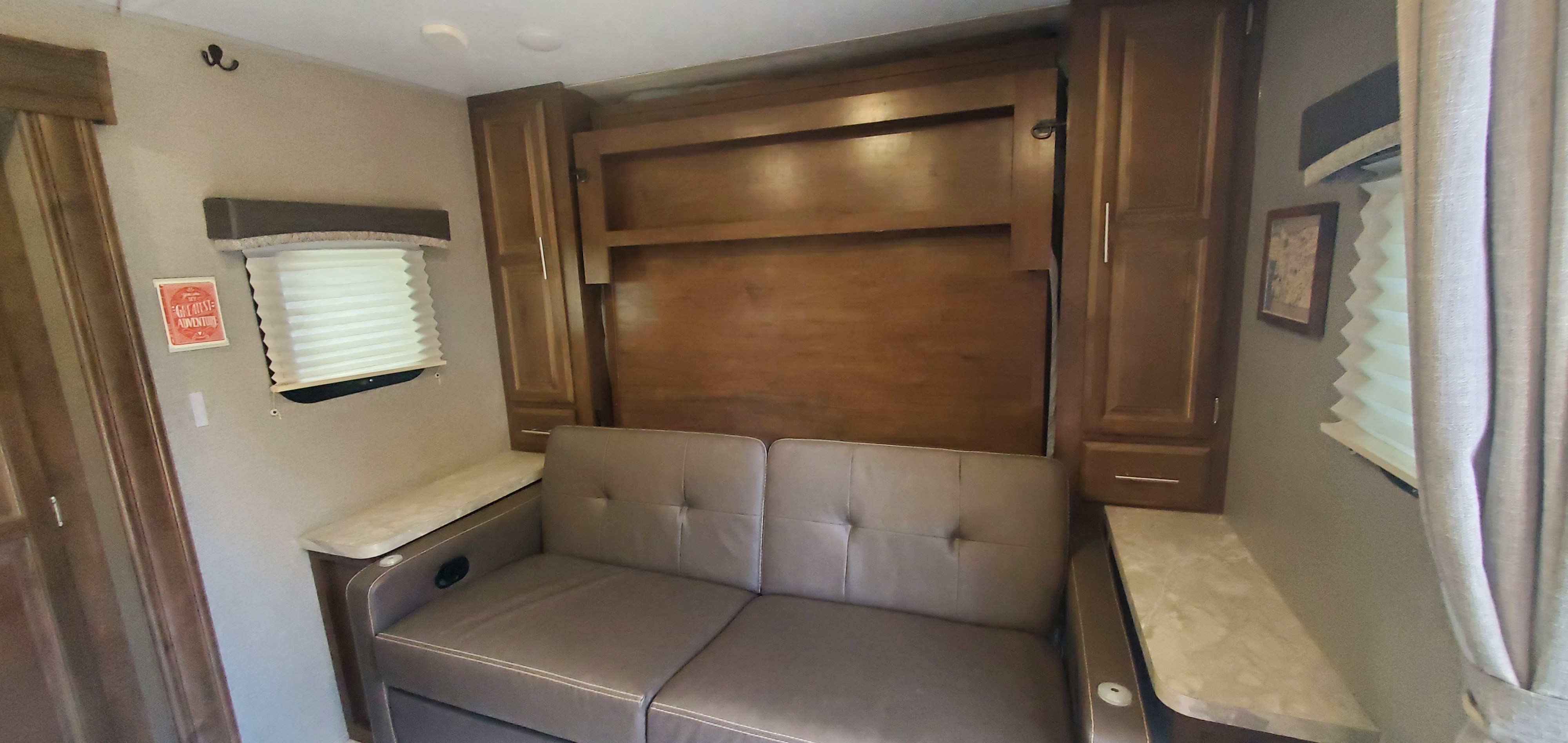 An RV rental living space featuring a sofa in front of a wall bed, built-in storage cabinets, and windows with blinds.. Forest River Rockwood Mini Lite 2019