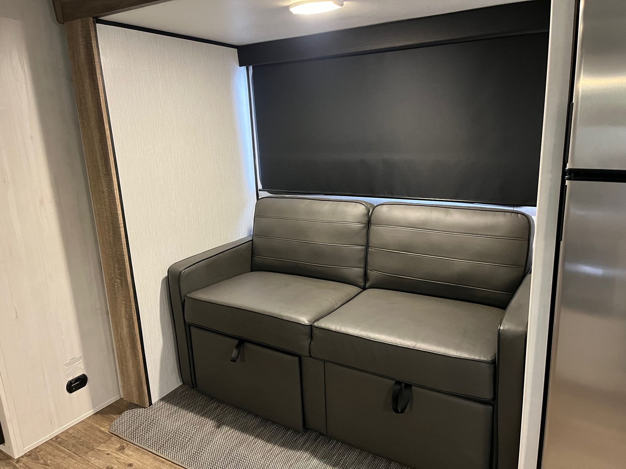 Interior of an RV rental showing a dark gray sofa with under-seat storage and a black window blind.. Heartland RVs Trail Runner 2022